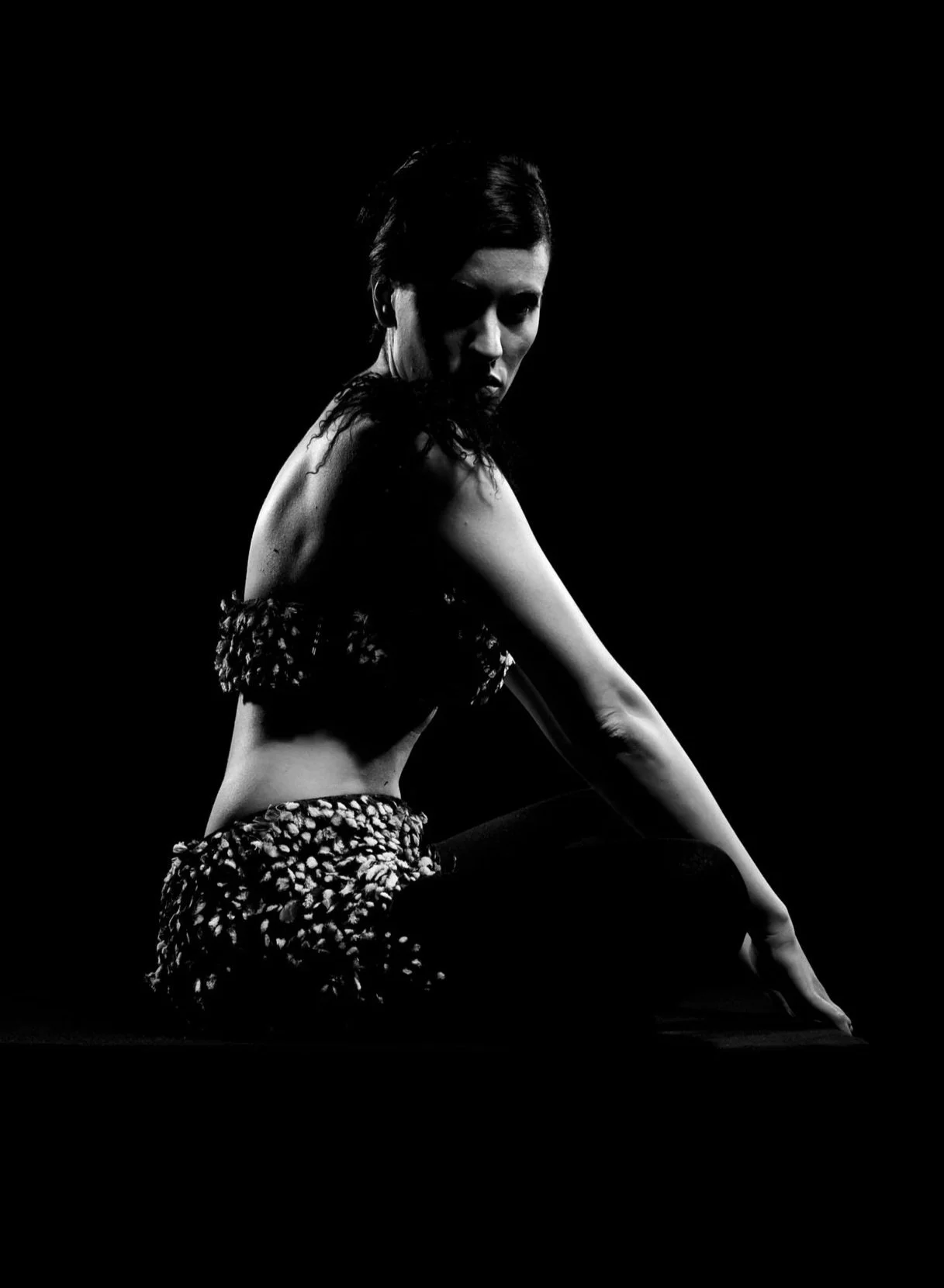 Black and white photo of a woman sitting with her left arm extended and looking over her shoulder against a black background.