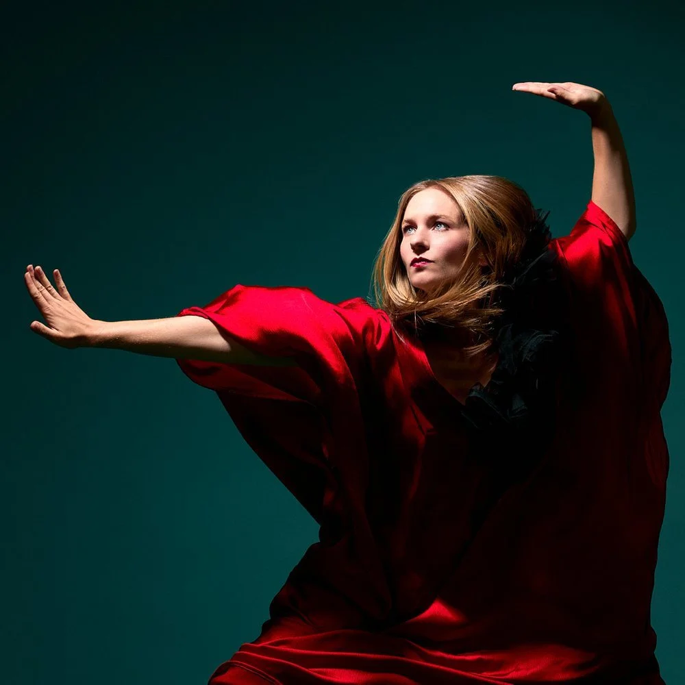 A woman with long hair in a red satin dress poses against a dark background, with one arm extended forward and the other raised above her head.