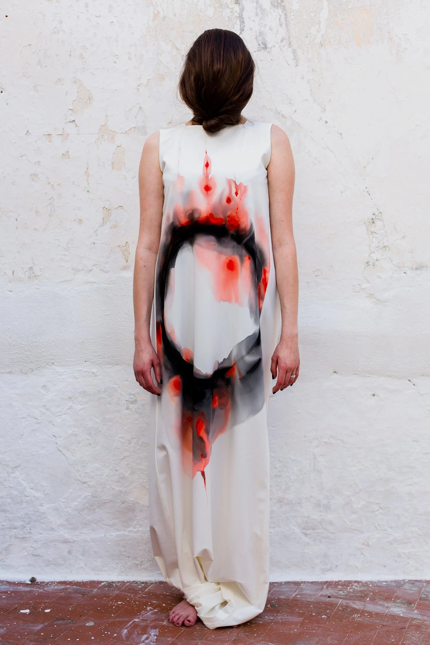 Woman standing against a white, textured wall, wearing a sleeveless white dress with black, red, and pink abstract paint splats.