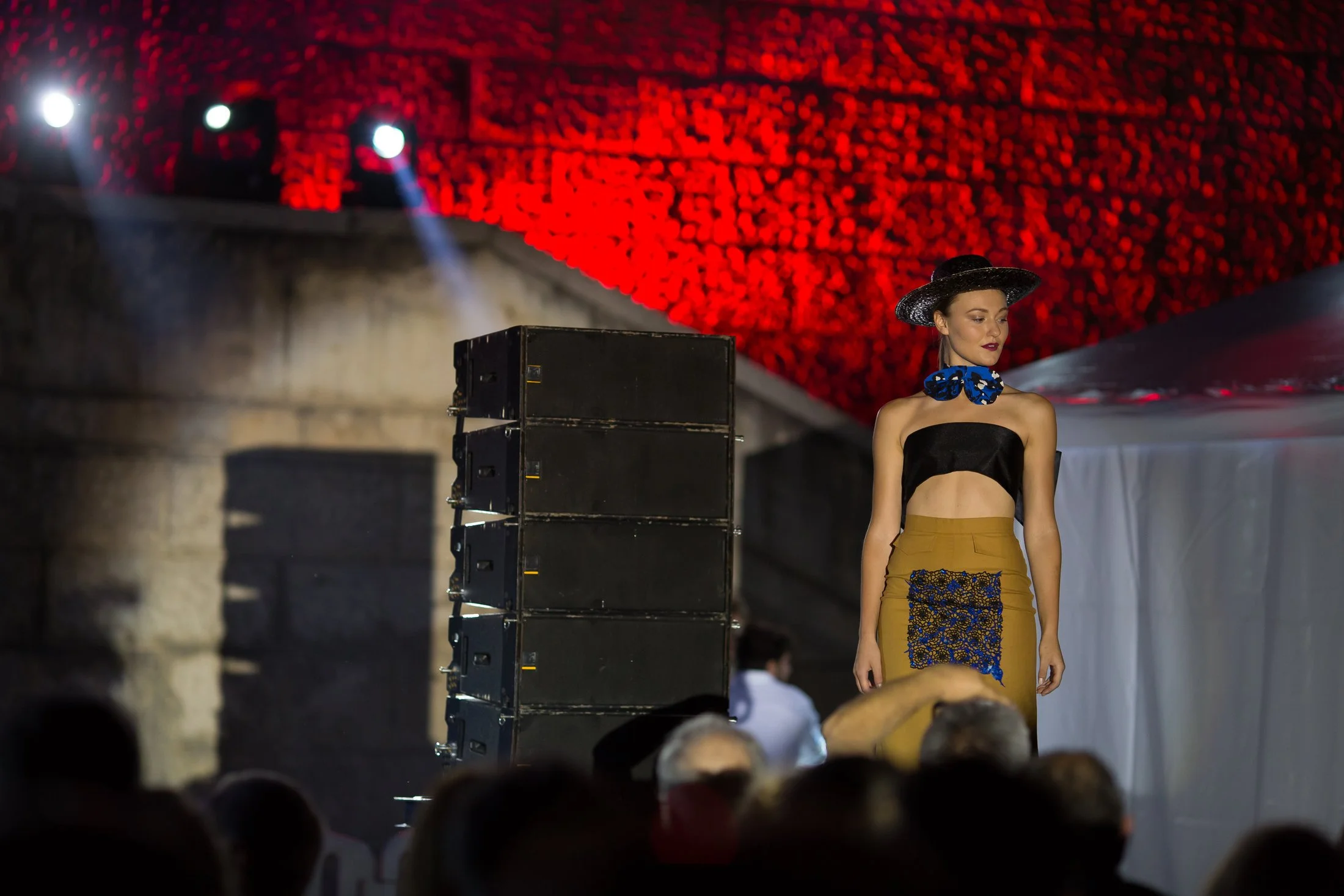 A woman walking on a runway during a fashion show with red artwork in the background and black speakers in the foreground.