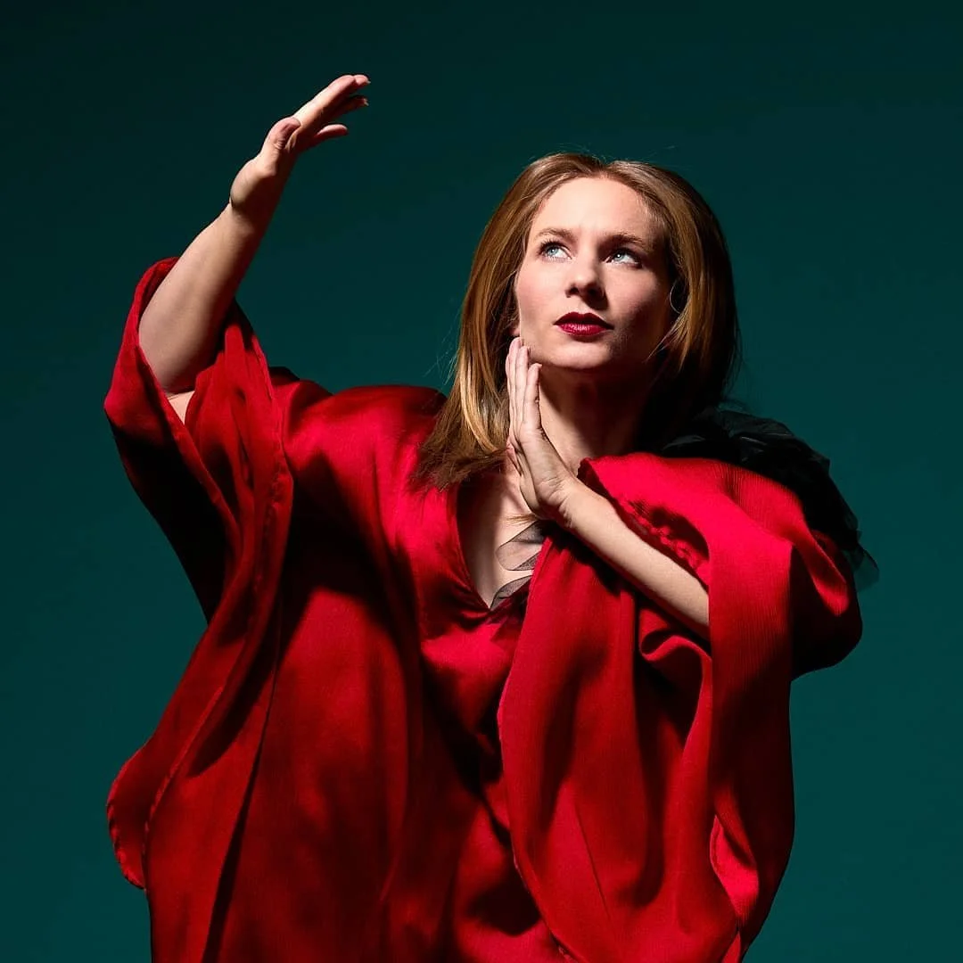 A woman with red hair wearing a red and black garment, striking a dramatic pose against a dark background.