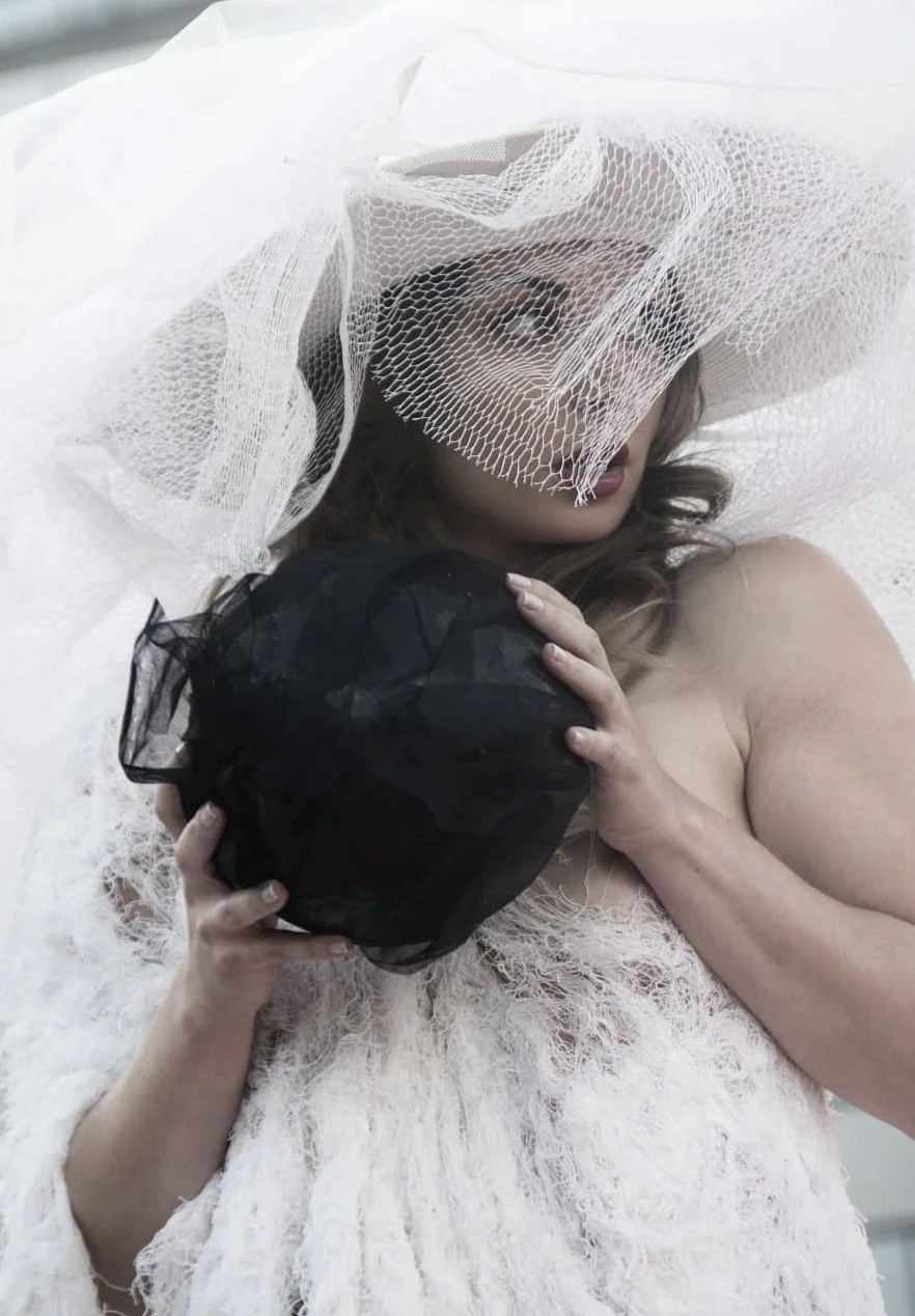 A woman wearing a large, white, mesh hat with a veil, holding a black object, and dressed in a fluffy white outfit.