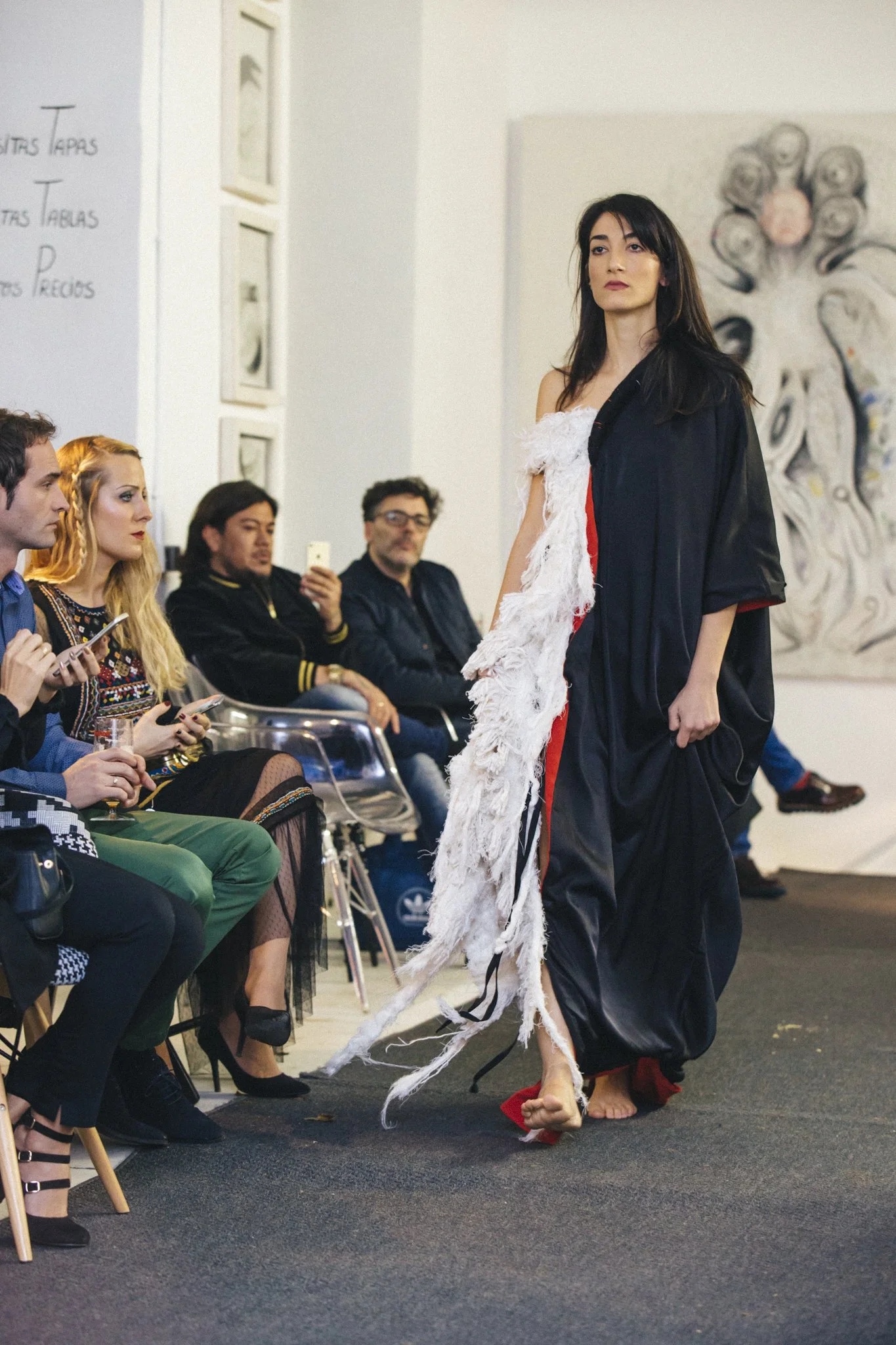 A woman with long dark hair is performing a traditional dance, barefoot, wearing a black and red garment with a white fringed accessory, as an audience watches attentively.