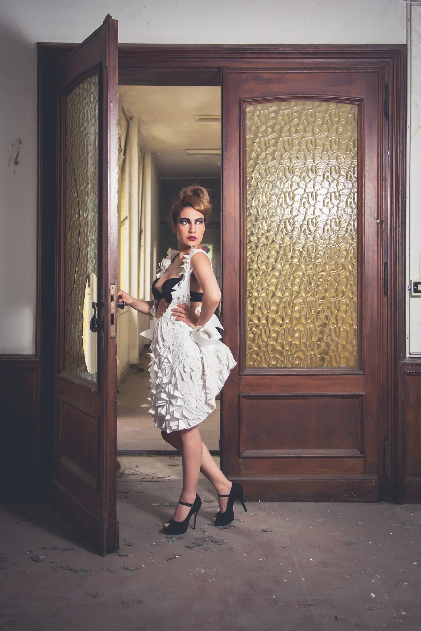 A woman with styled hair in an updo, wearing a black and white dress with a textured, sculptural skirt, and black high heels, stands in an abandoned building doorway with wood and textured glass panels.