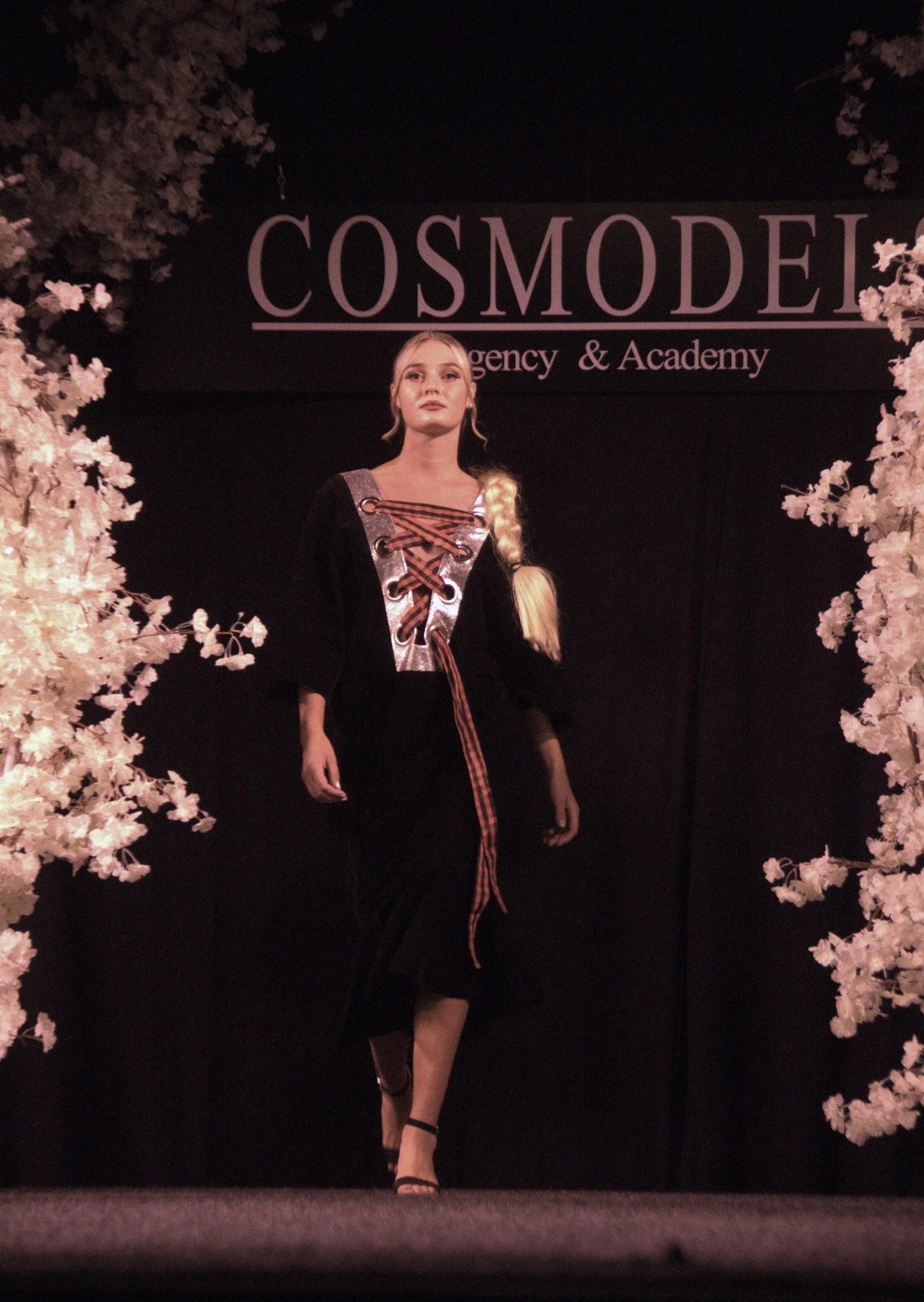 A fashion model walking on stage at a runway show, with a large sign reading 'COSMODEL' and 'Agency & Academy' behind her, framed by white floral decorations.