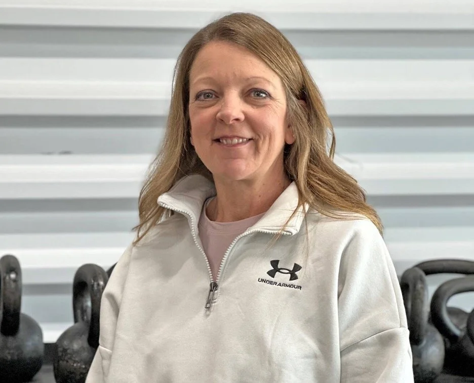 A smiling woman with light brown hair in a ponytail, wearing a white Under Armour zip-up jacket, standing in front of kettlebells in a gym.