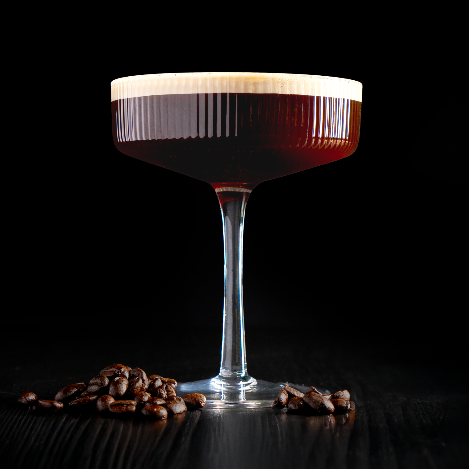 A martini glass of coffee cocktail with a layer of cream on top, surrounded by coffee beans on a dark surface against a black background.