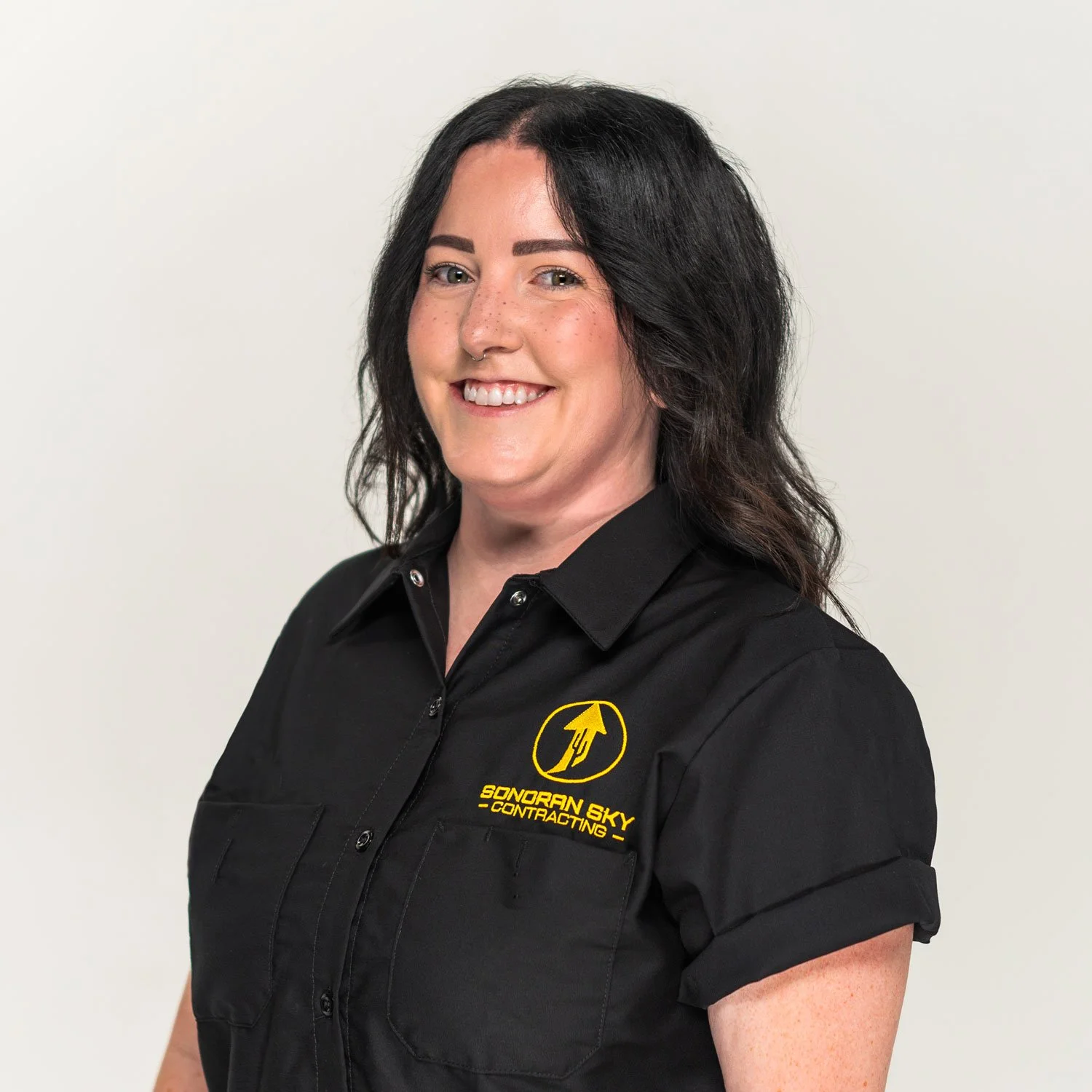A woman with dark, wavy hair, smiling, wearing a black shirt with a yellow logo.
