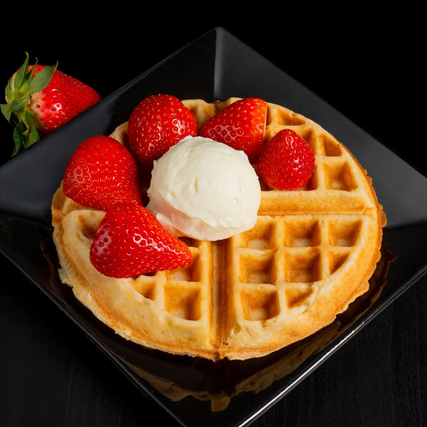 A golden-brown waffle topped with strawberries and a scoop of vanilla ice cream on a black plate.