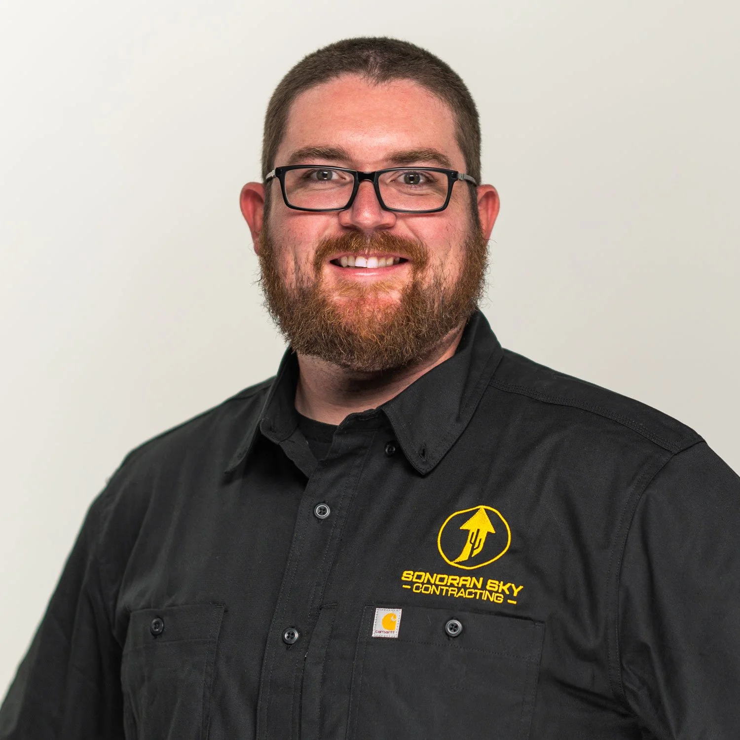 A man with glasses and a beard, smiling, wearing a black shirt with a yellow logo, against a plain white background.
