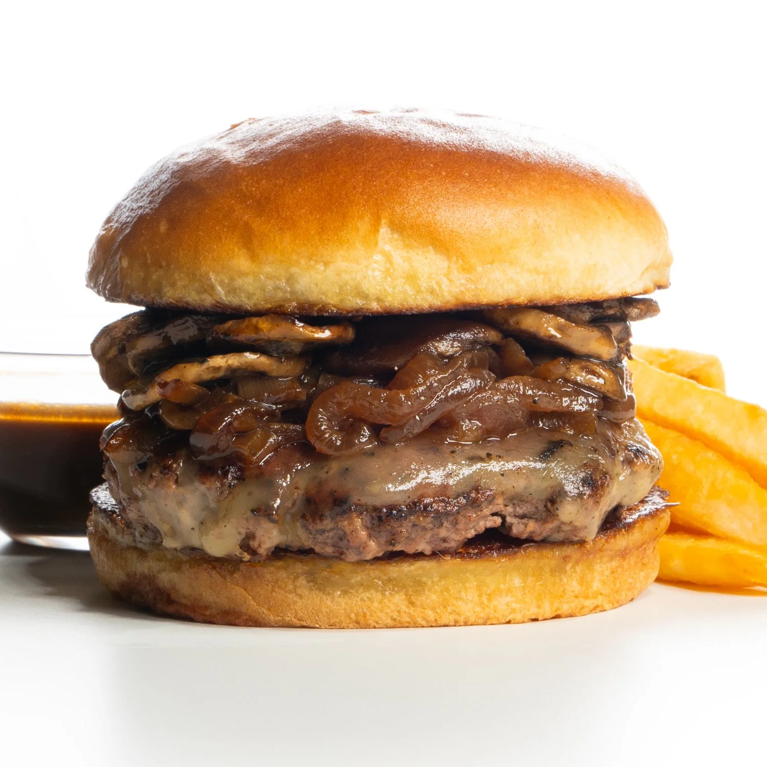 Close-up of a French onion cheeseburger with caramelized onions, melted cheese, and a toasted bun, served with a side of French fries and a small bowl of dipping sauce.