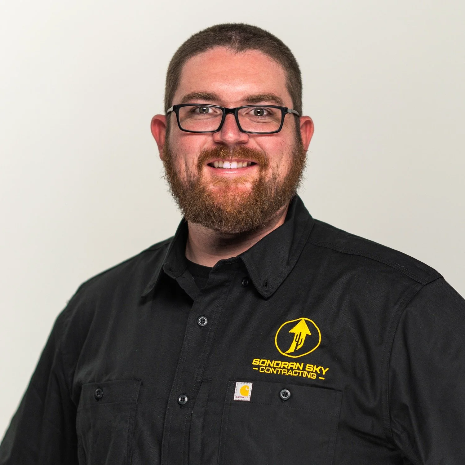 A man with glasses and a beard, smiling, wearing a black collared shirt with a yellow logo.