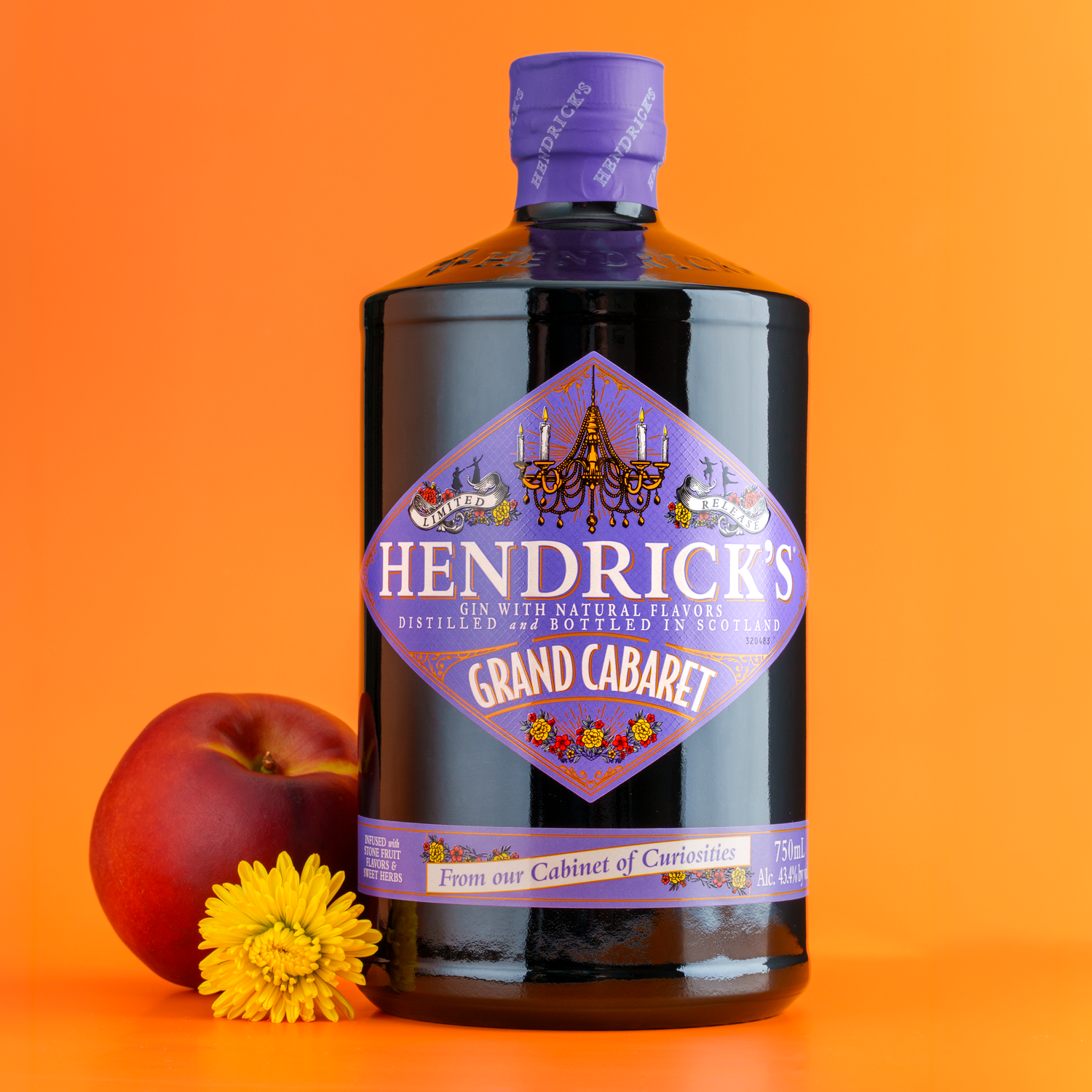 A bottle of Hendrick's Grand Cabaret gin with a purple label and cap, placed against an orange background. Next to the bottle, there is a red peach and a yellow flower.