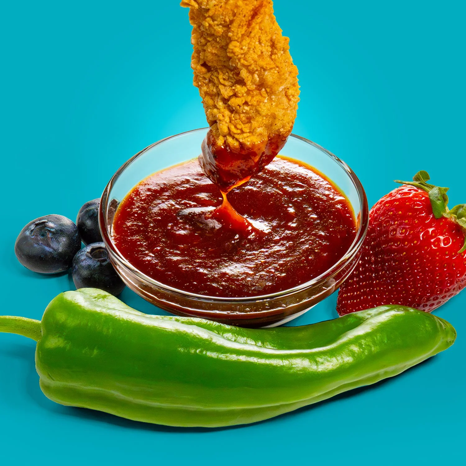Fried chicken strip dipped in barbecue sauce in a small glass bowl, surrounded by blueberries, a strawberry, and a green bell pepper on a blue background.