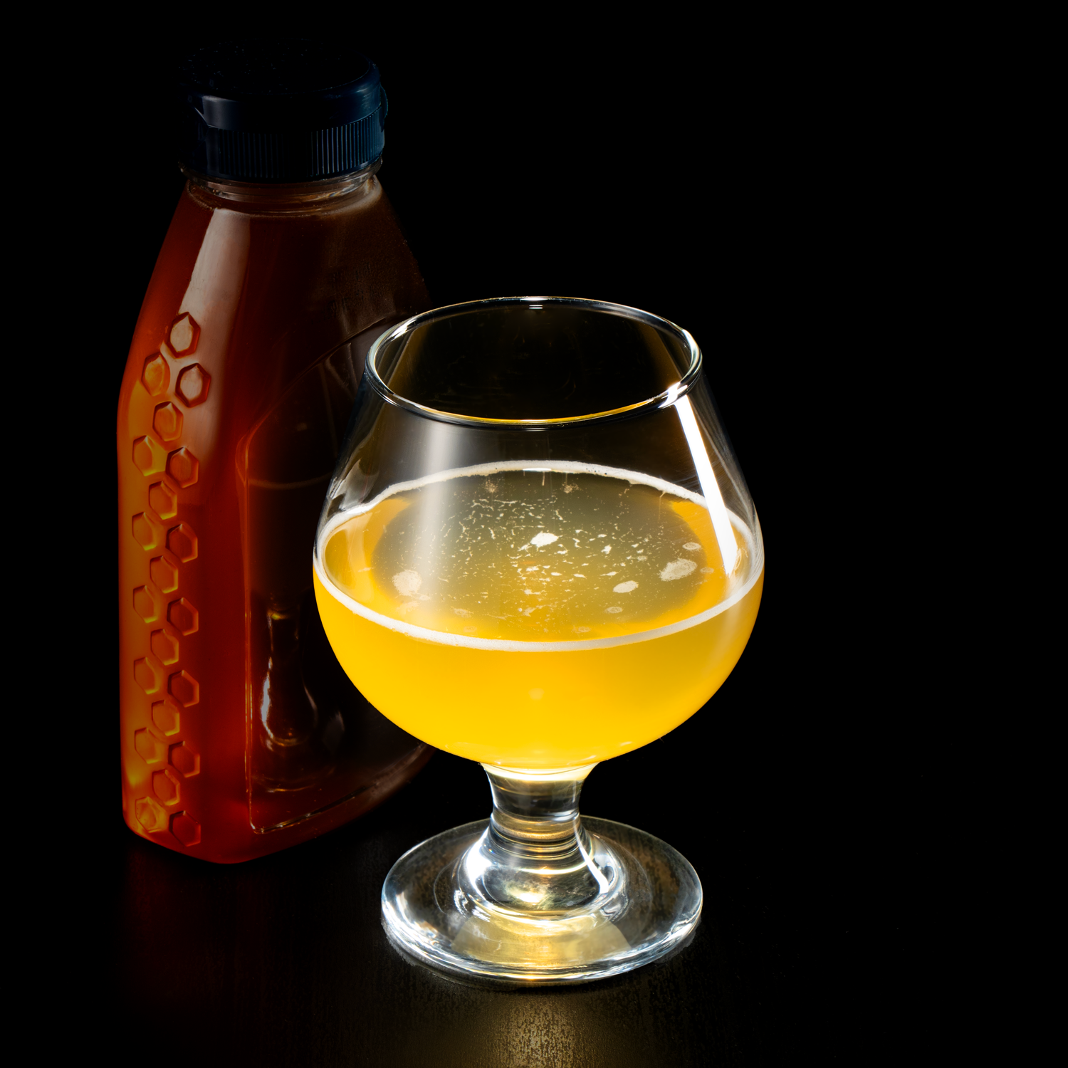 A glass of yellow mead with foam on top, placed on a black surface with a dark background. A bottle of honey is positioned behind the glass.
