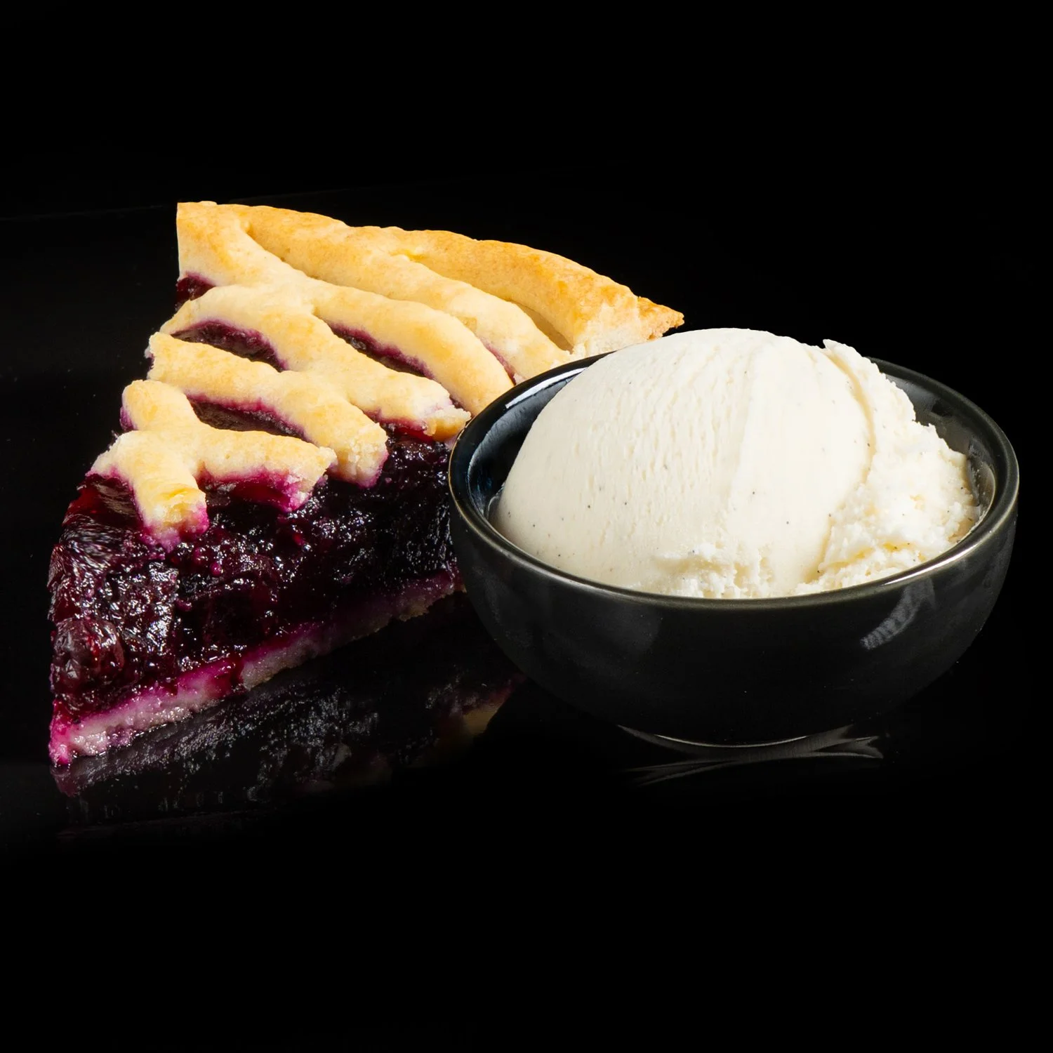 Slice of blueberry pie with lattice crust and scoop of vanilla ice cream in a black bowl on a black background.