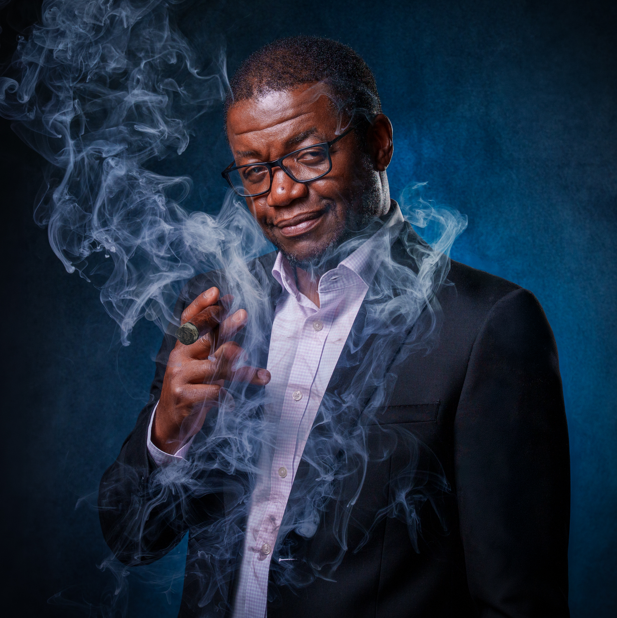 A man with glasses in a suit smokes a cigar with smoke swirling around him against a dark blue background.