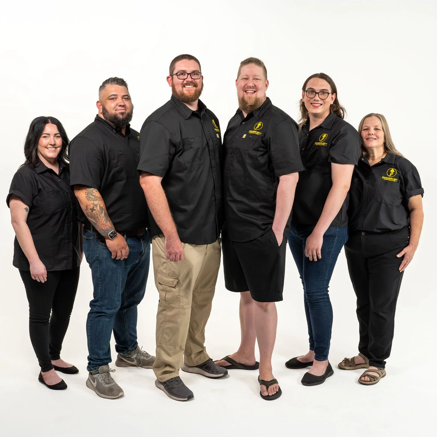 Group of six people smiling and standing together, dressed in black shirts with yellow logos, against a plain white background.
