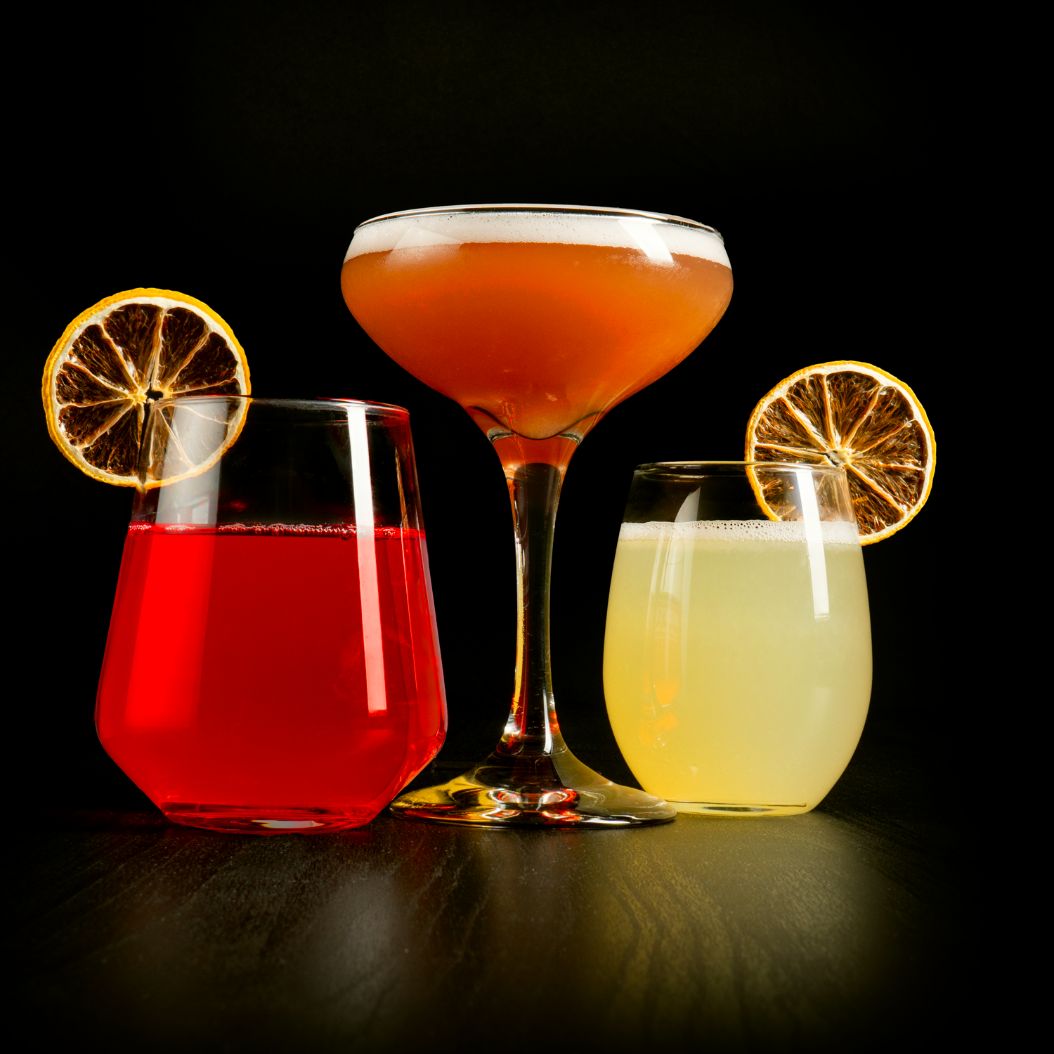 Three colorful cocktails with dried orange slices garnishes on a dark background.