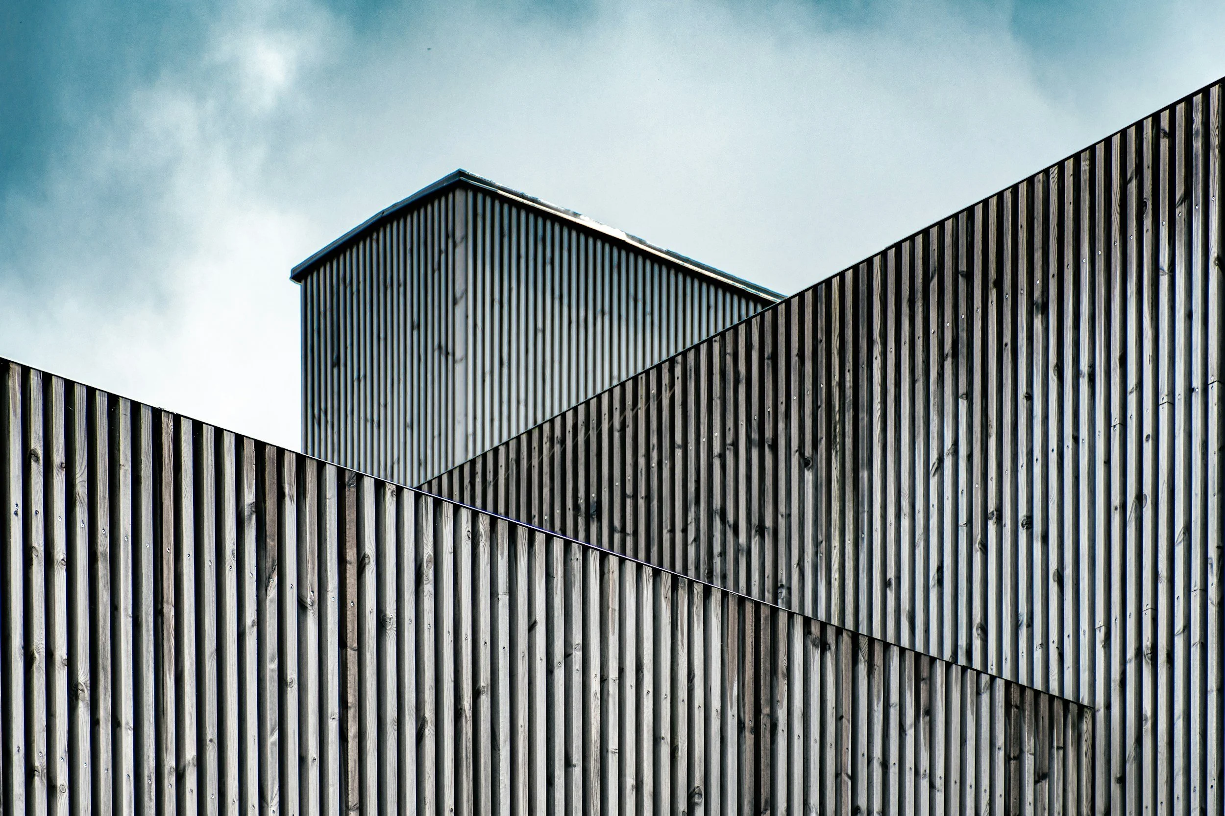 Close-up of modern building with vertical wooden slats on facade, blue sky with clouds in background.
