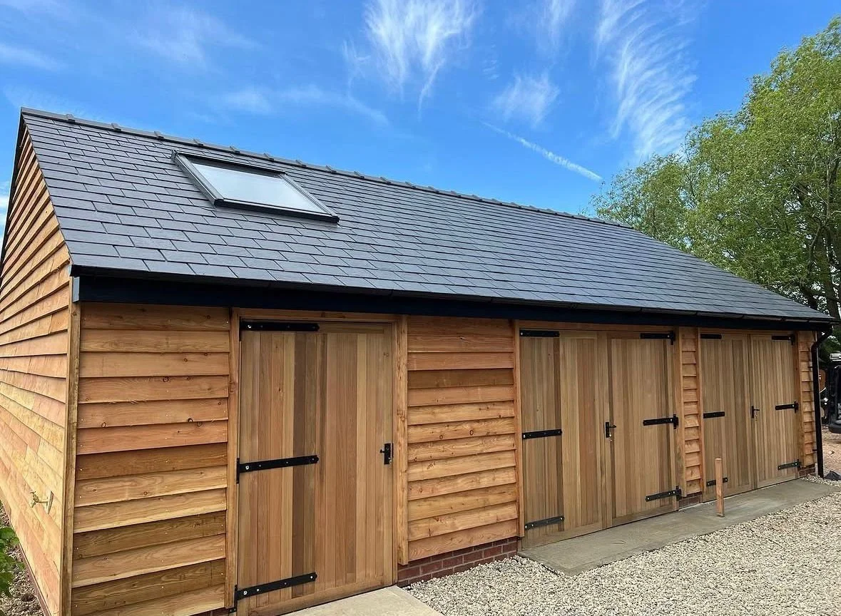 A wooden garage with four sliding barn doors and a black shingle roof, with a skylight on the roof, set outdoors with trees and a blue sky in the background.