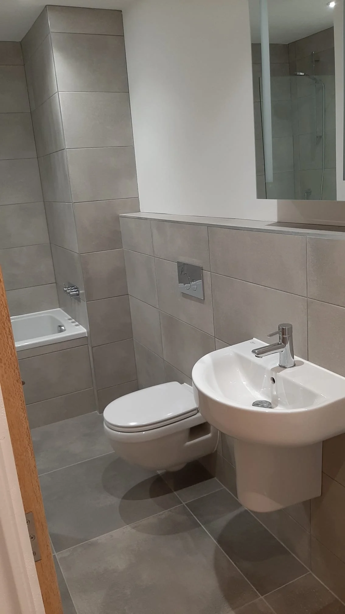 A small, modern bathroom with gray tiled walls and floor, featuring a white toilet, a white sink with a silver faucet, a rectangular mirror, and a walk-in shower with glass doors in the background.