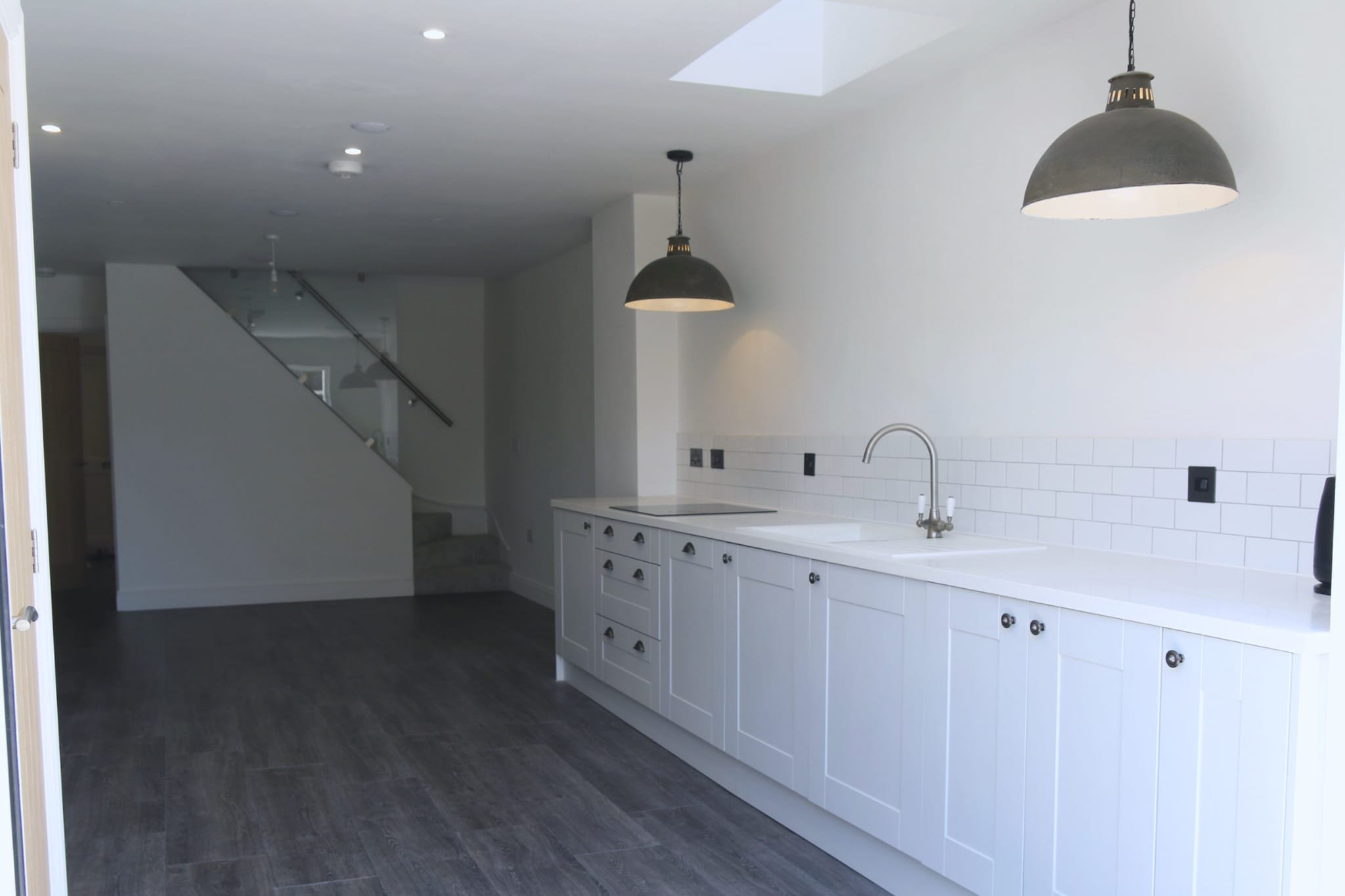 Modern white kitchen with black pendant lights, dark wood flooring, white cabinets with black handles, and a white subway tile backsplash.