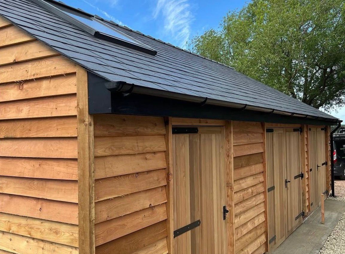 A wooden outdoor shed with a sloped black shingle roof, three wooden doors with black hardware, and a gravel area in front during daytime with a clear blue sky and green trees in the background.