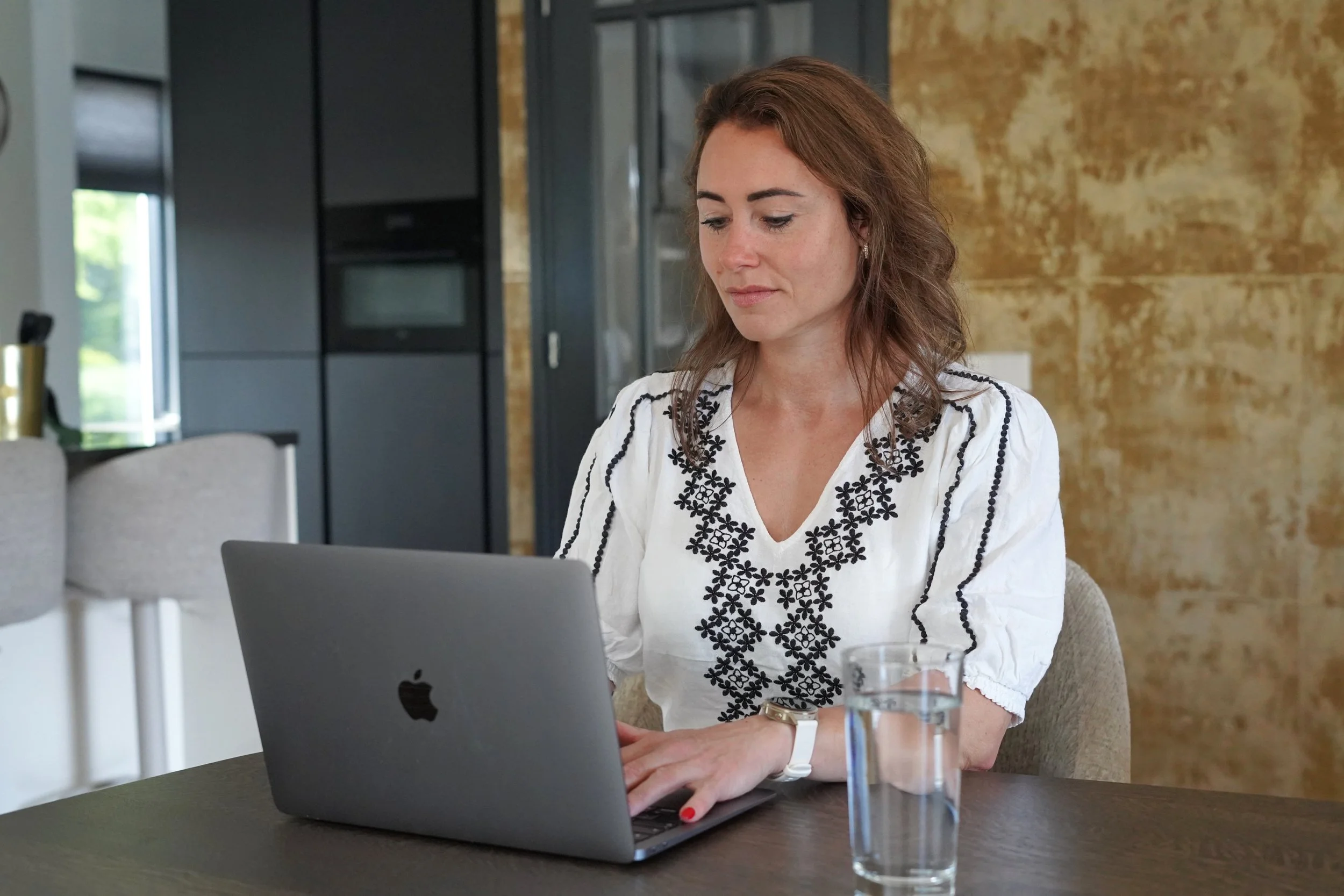 Vrouw werkt op een laptop in een huiselijke omgeving, met een glas water op tafel.