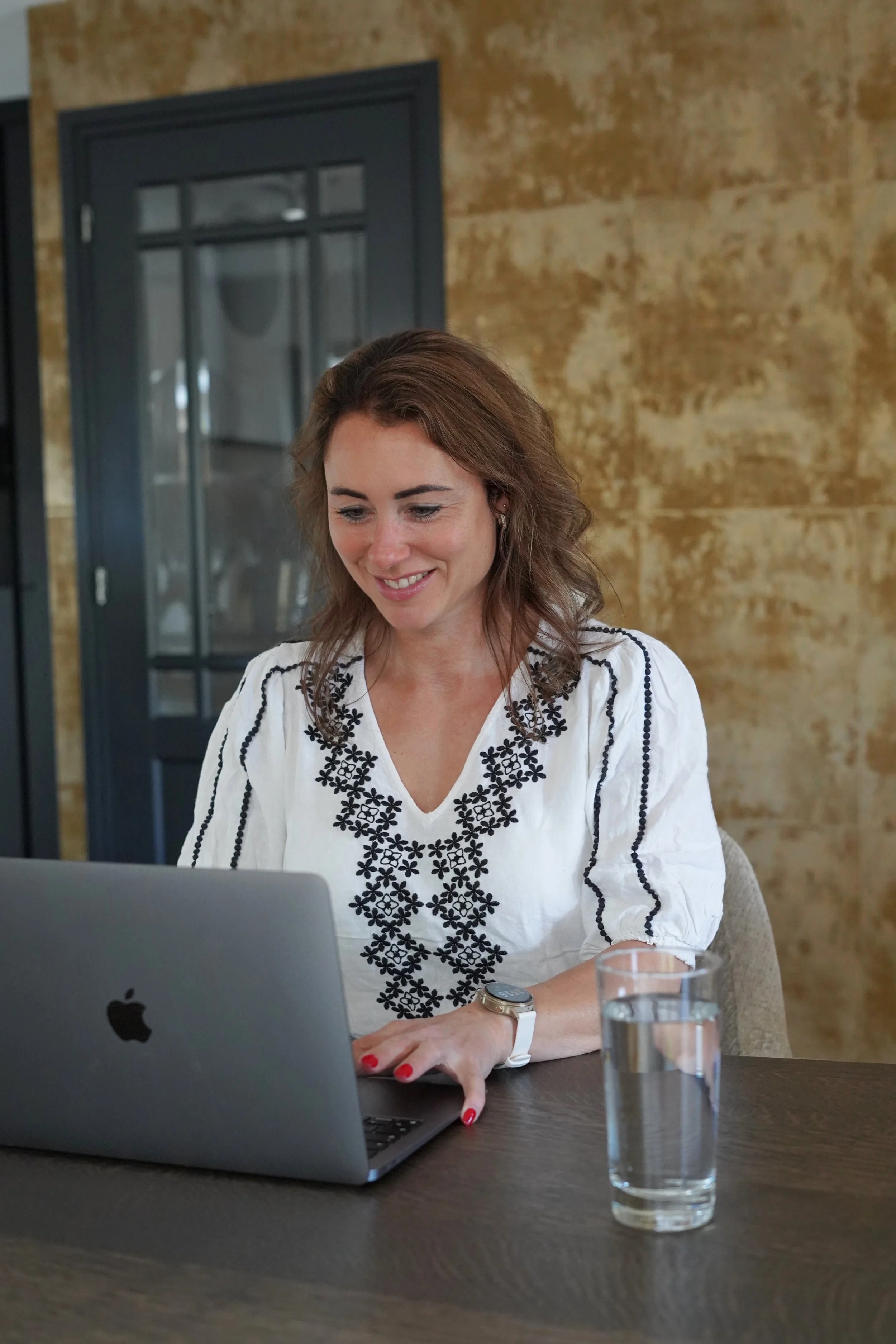 Vrouw zit aan een tafel met een open MacBook, draagt een witte blouse met zwarte borduursels, heeft bruin halflang haar, en een horloge aan haar linkerpols. In de glas is water, op de tafel.