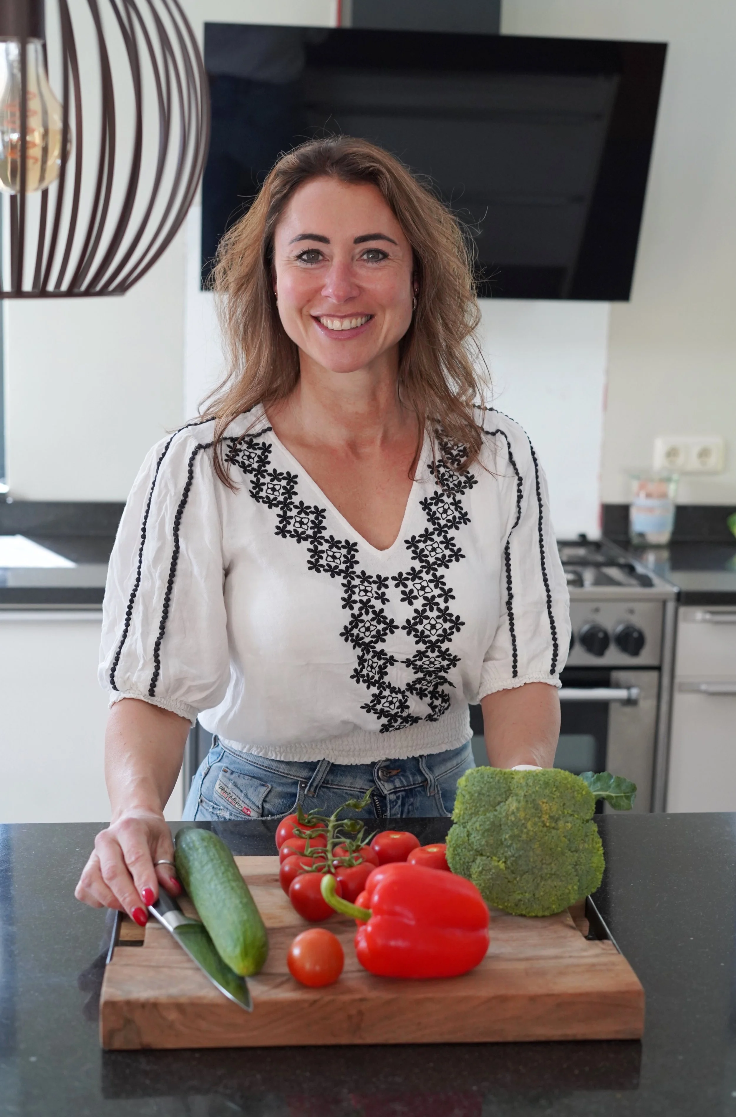 Een vrouw staat in een keuken met een snijplank vol verse groenten, waaronder tomaten, komkommer, broccoli en paprika, klaar om te koken.