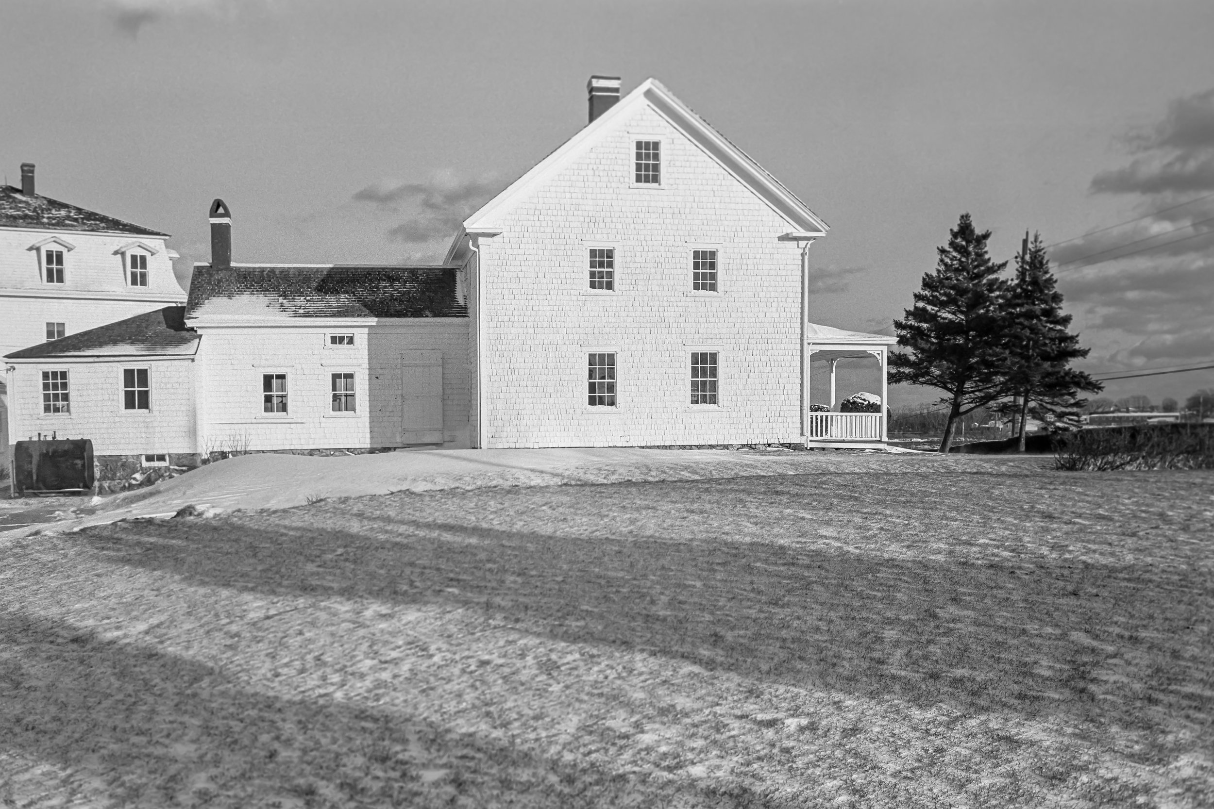 A black-and-white photo of a large white house with multiple floors and windows, a porch on the right side, a snow-covered yard, a tree, and a cloudy sky.