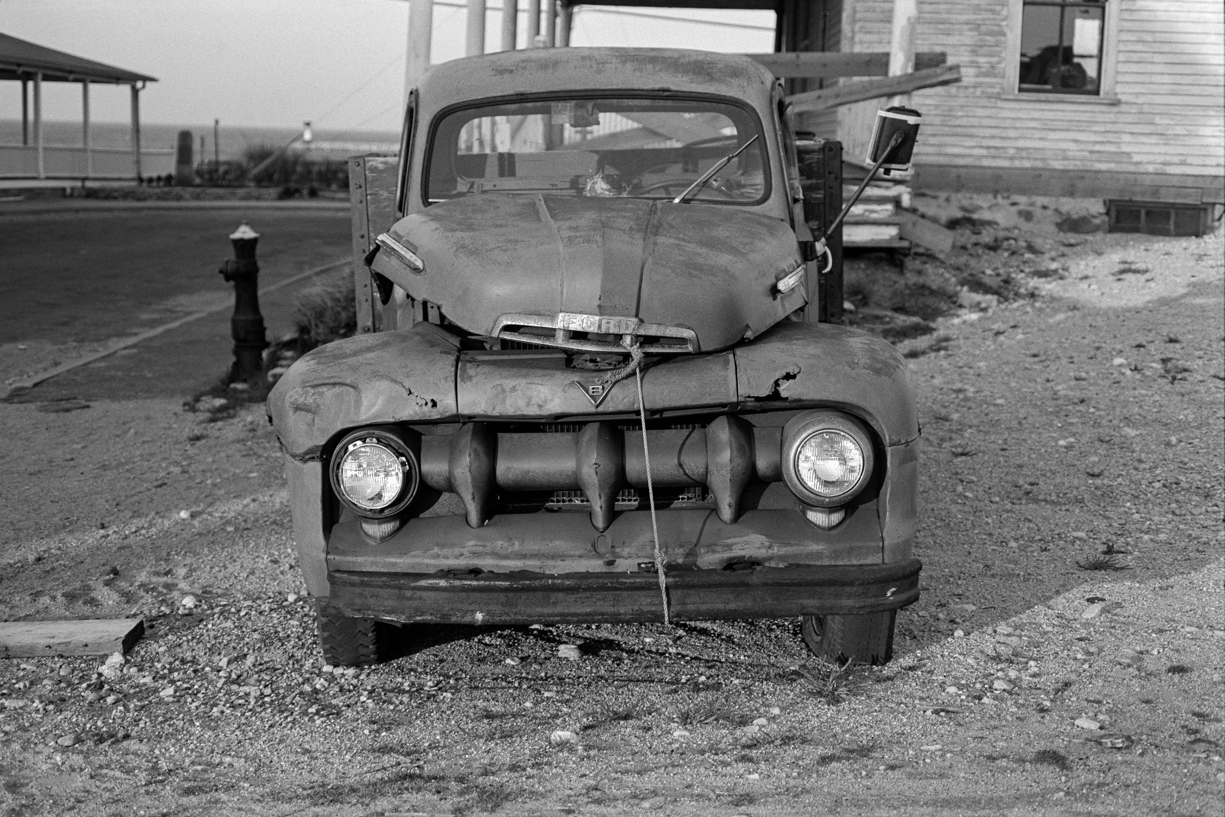 An old broken-down Ford pickup truck with a rusty and damaged front hood and missing parts, parked in a rural area in front of a wooden house. The photo is in black and white.