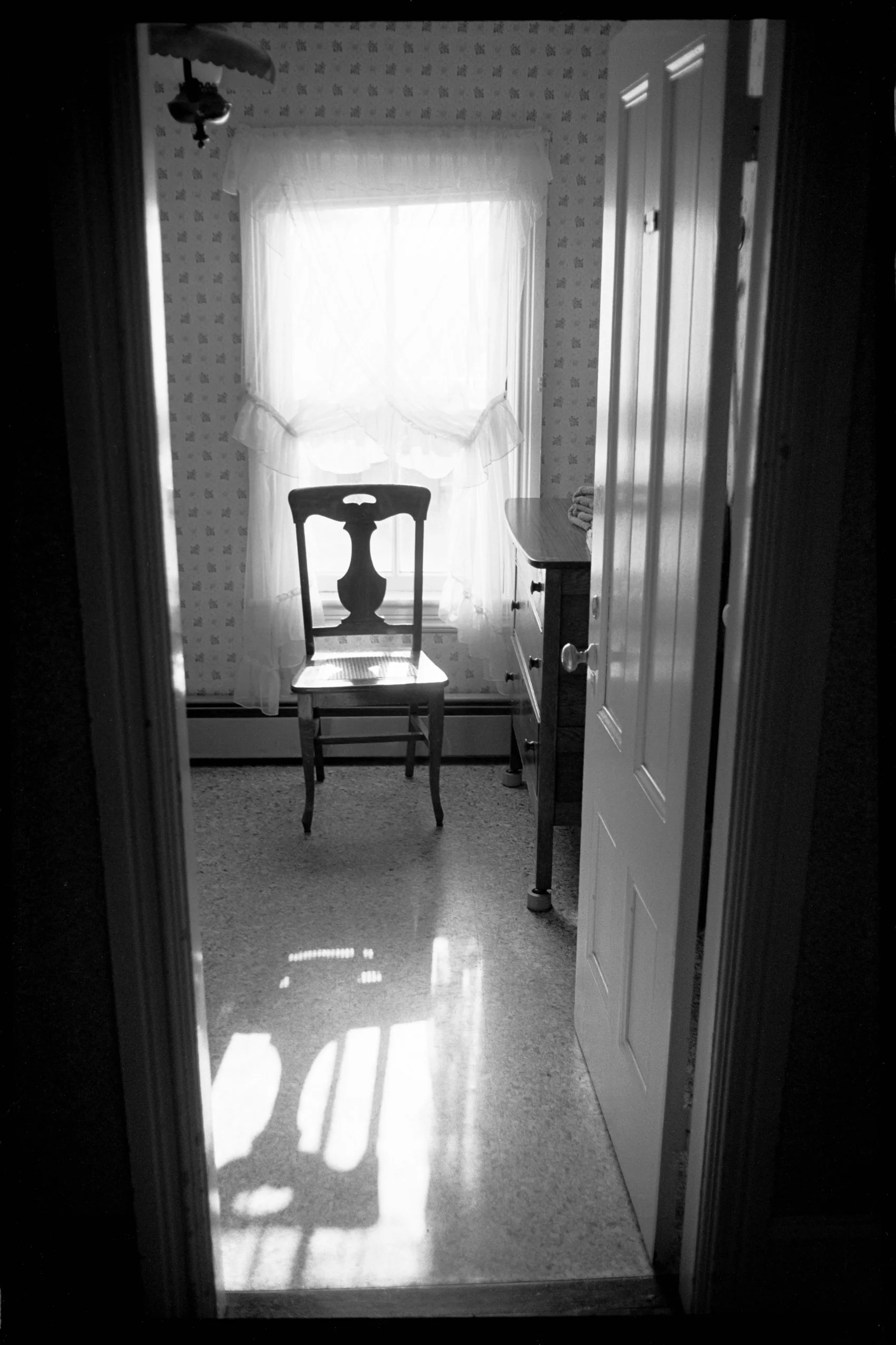 A doorway opening into a small room with a chair facing a window, a cabinet with folded towels, and a door on the right. Sunlight filters through the window, casting shadows on the floor.