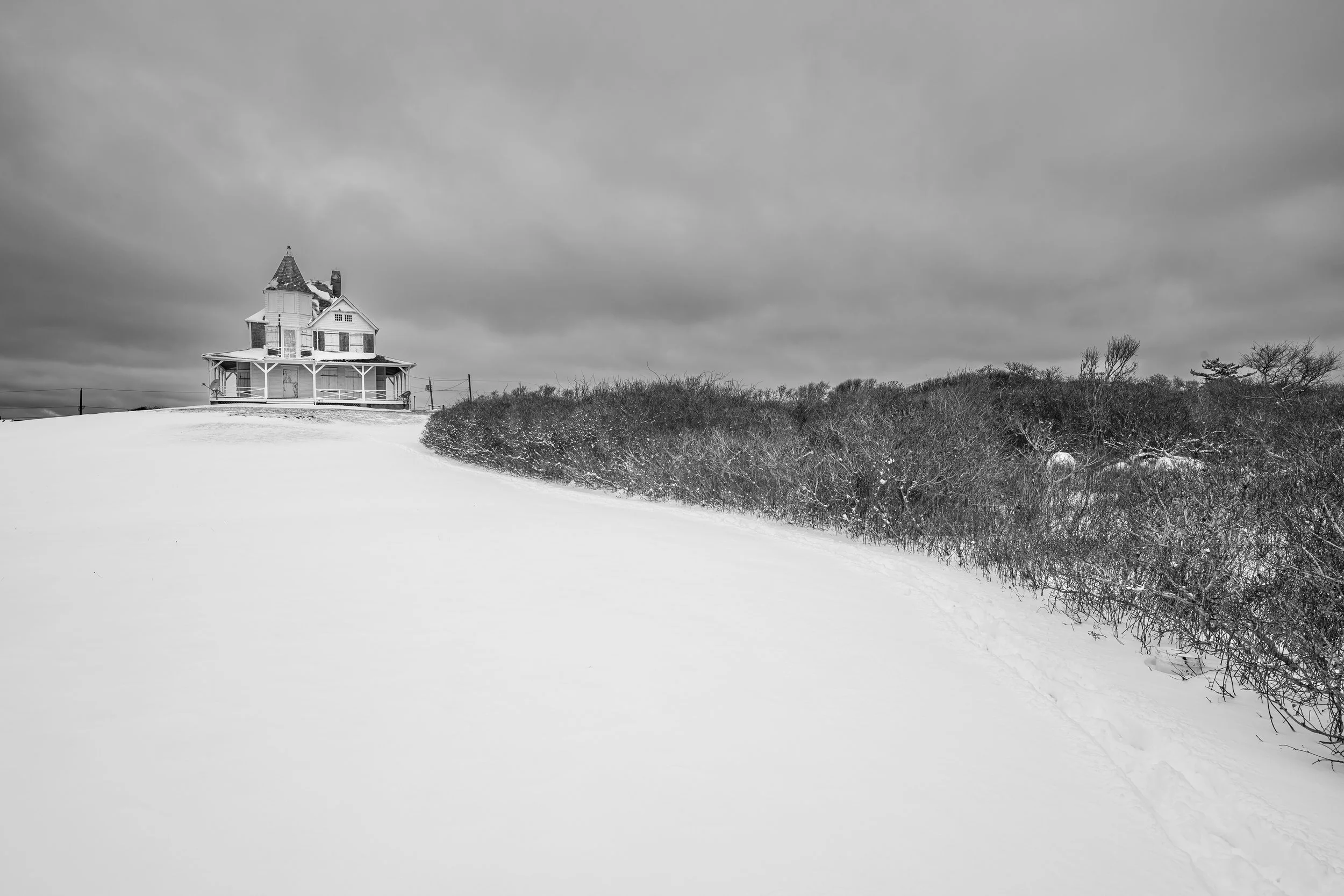 A black and white photograph of a snow-covered landscape with a house on a hill, bushes, and a cloudy sky.