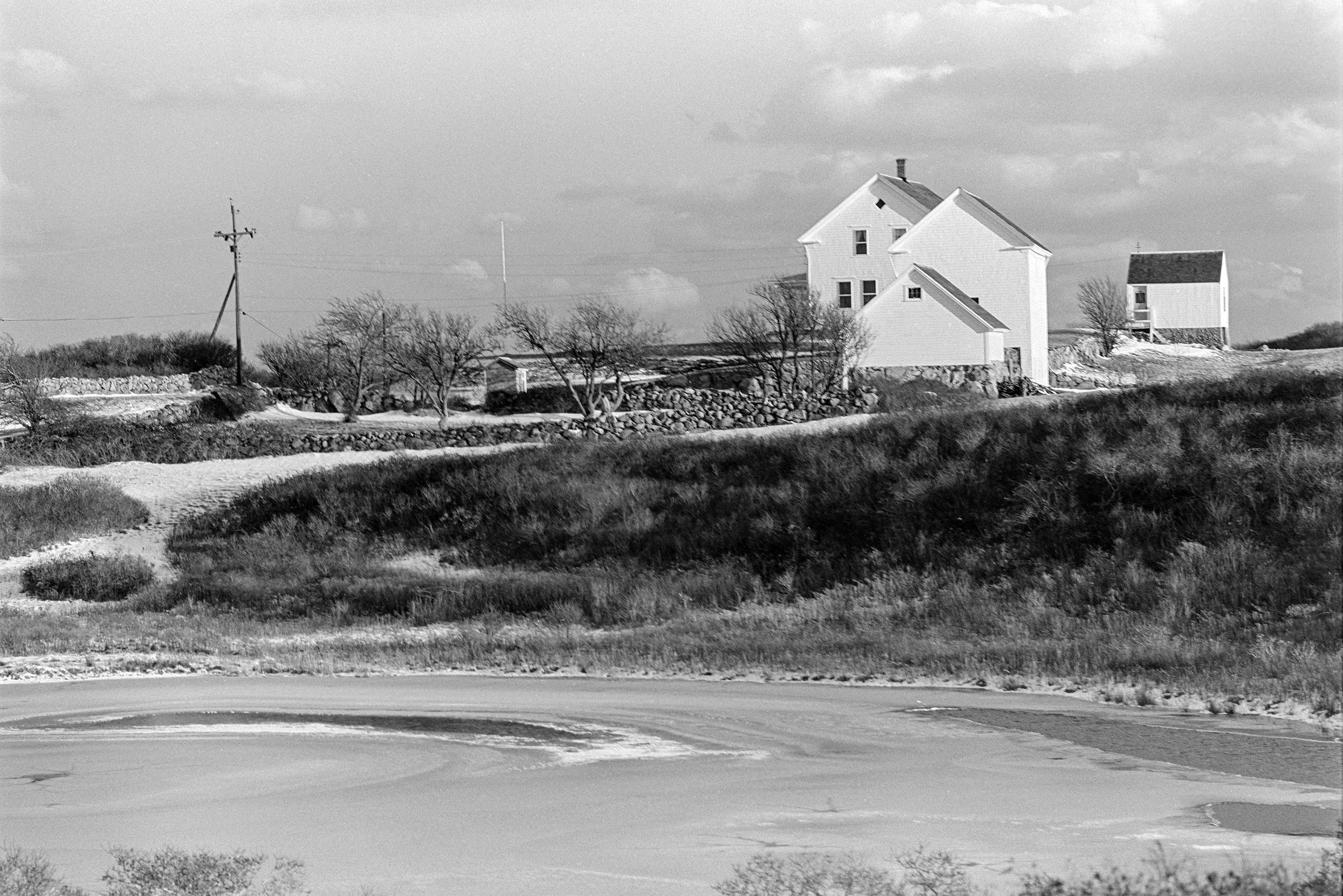Black and white photo of a rural landscape with a winding road, trees, a house, and power lines.