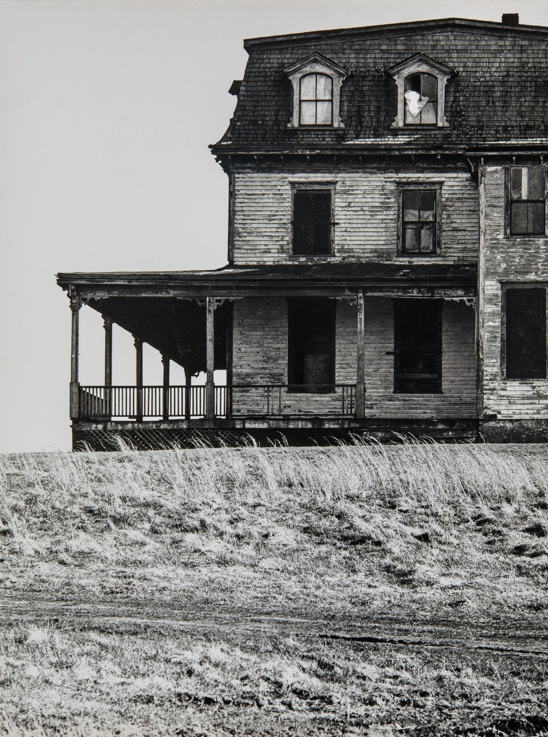 A black and white photograph of an old, weathered wooden house with a porch, situated outdoors on grassy land.