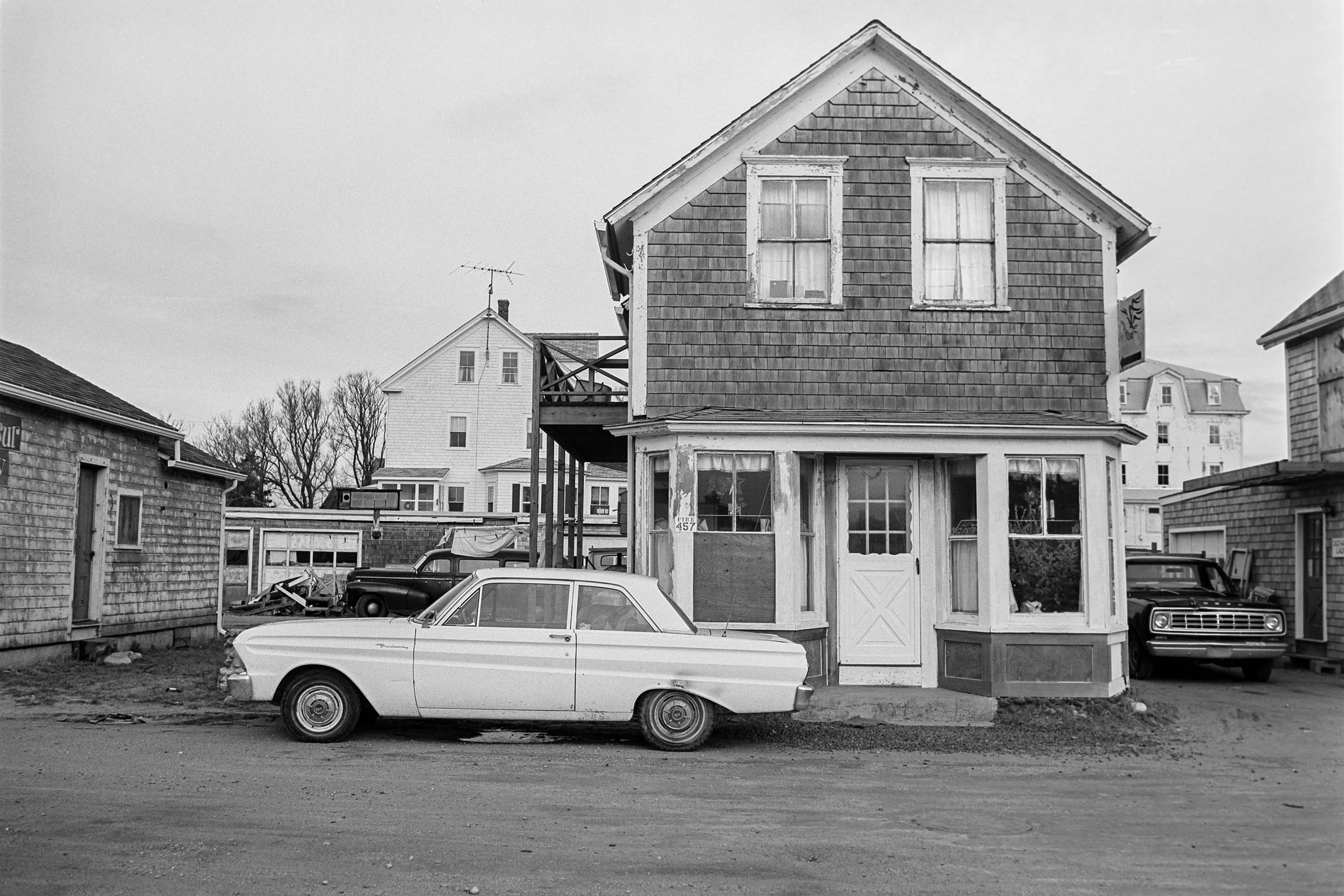A black and white photograph of an old house with weathered siding and peeling paint, parked cars including a vintage white car in the foreground, and other houses in the background
