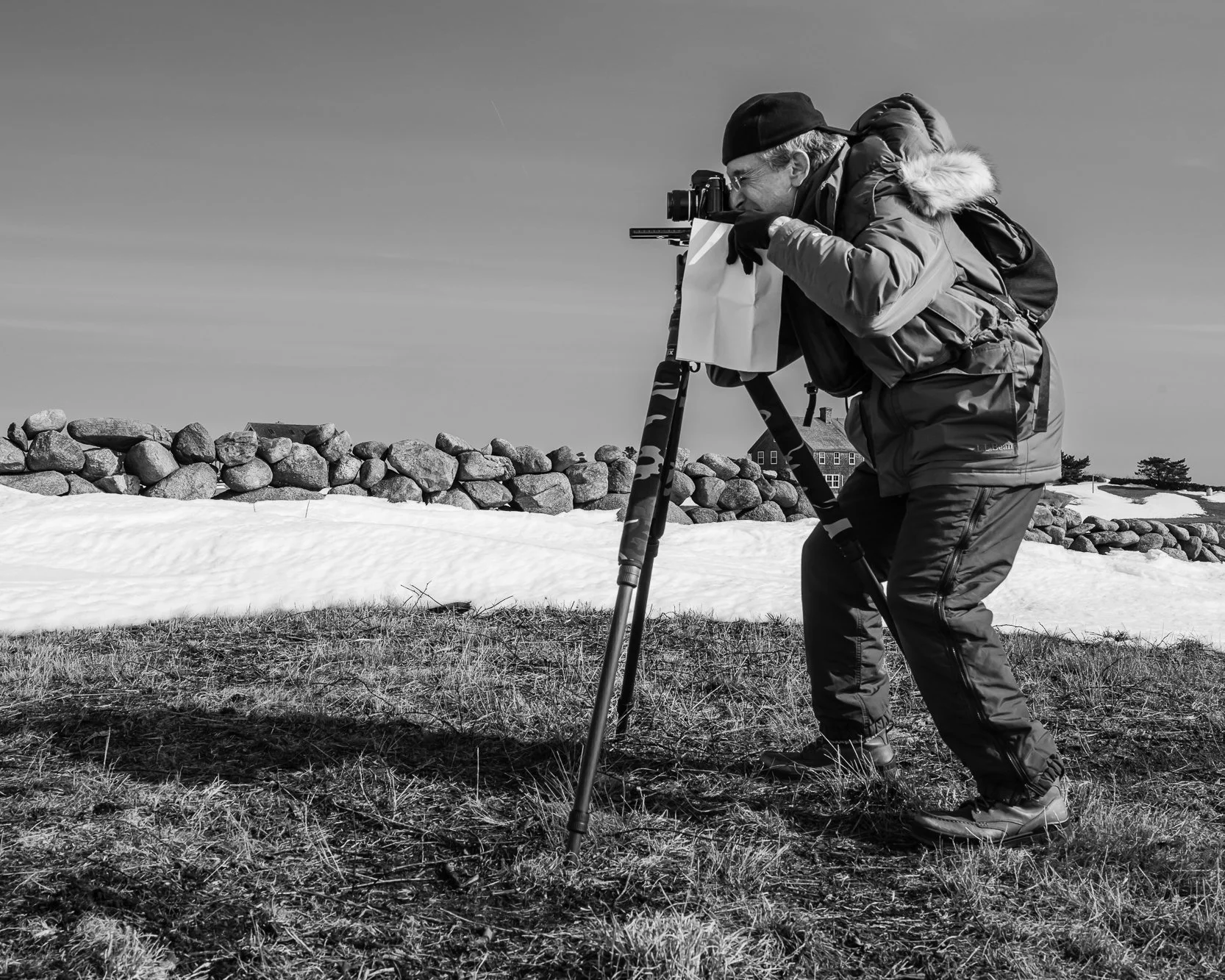 A man wearing winter gear, including a hat and gloves, is using a transit level or surveying instrument outdoors on a snow-covered area with grass visible in the foreground. There is a stone wall and a house in the background.