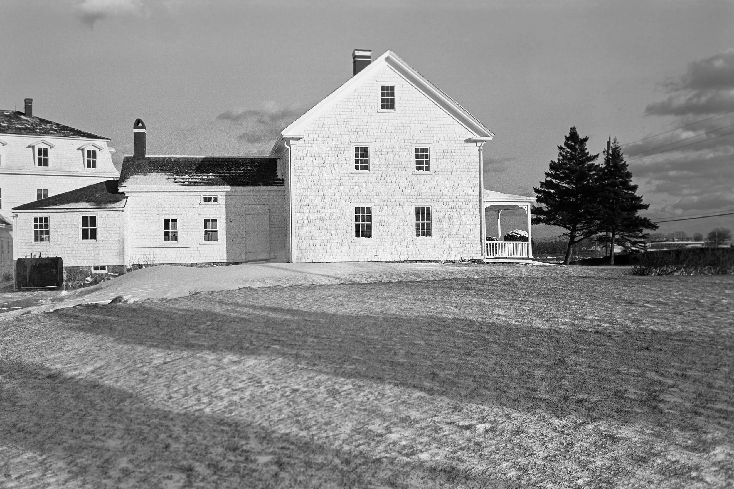 A black and white photo of a house with a steep gable roof, several windows, and a porch, situated in a rural area with trees and open land, and some snow on the ground.