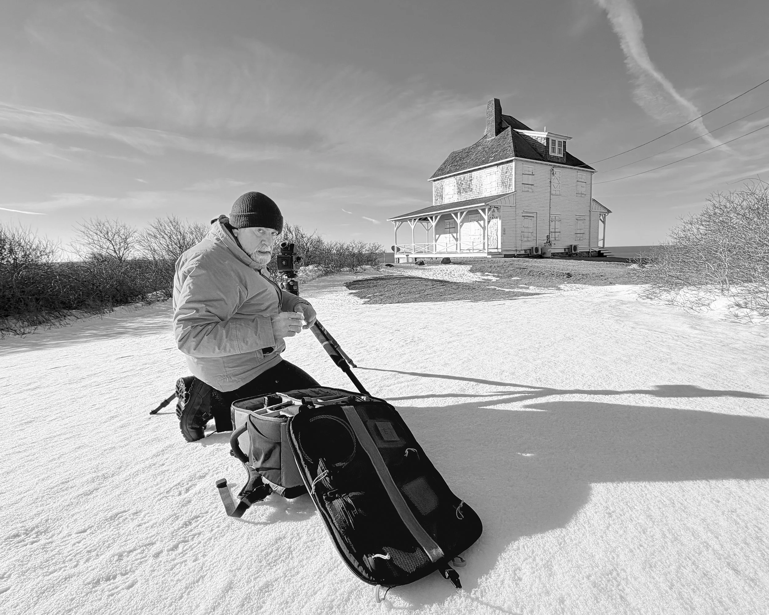A person dressed warmly with a beanie and jacket kneeling on snow-covered ground, holding a camera mounted on a tripod, with a house and leafless trees in the background on a clear day.
