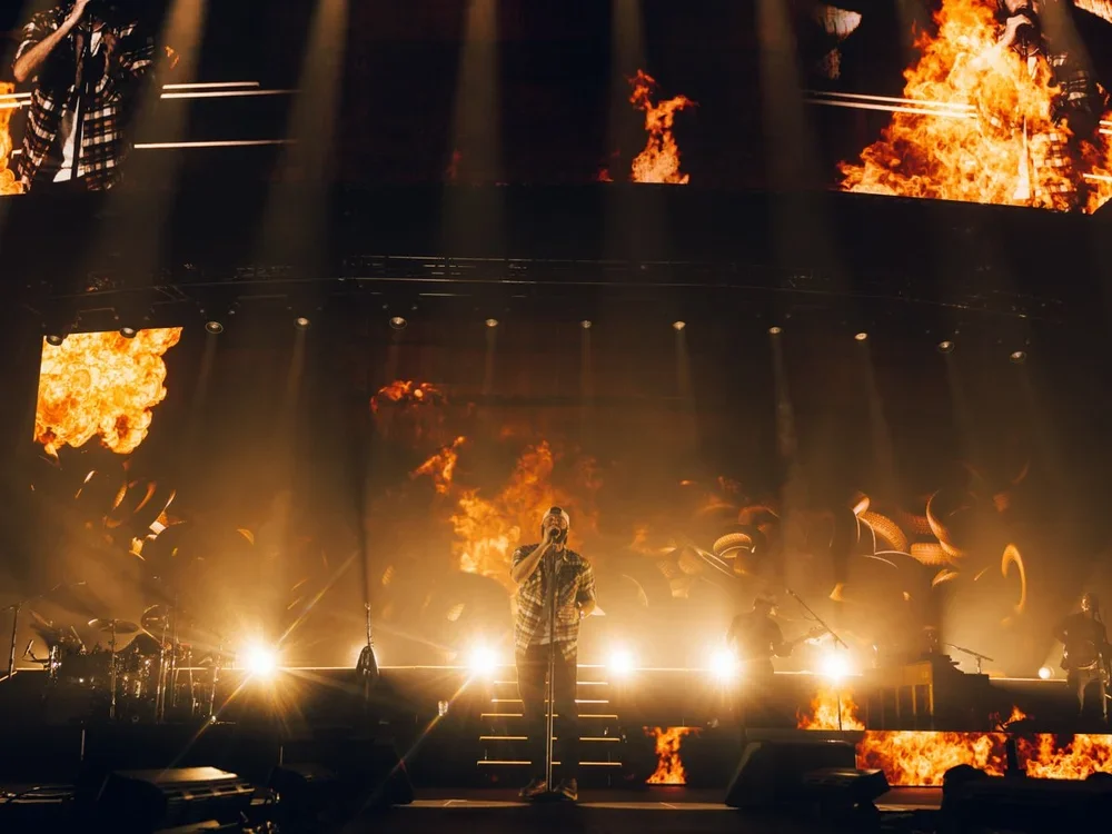 An artist performing on stage with bright lights and fiery visuals projected behind him, creating a dramatic concert atmosphere.