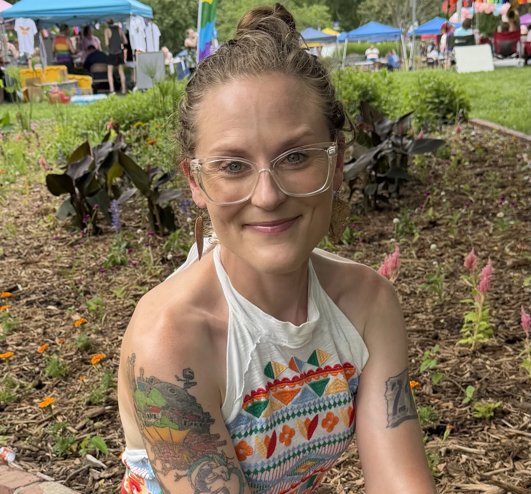 A woman with glasses and tattoos on her arm smiling at the camera, outdoors at a festival with colorful tents, trees, flowers, and people in the background.