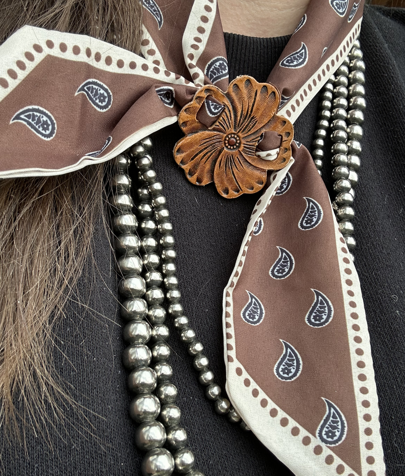 Close-up of a person wearing a brown bandana with a paisley pattern, a large leather flower brooch, and pearl necklaces. The person is dressed in black.
