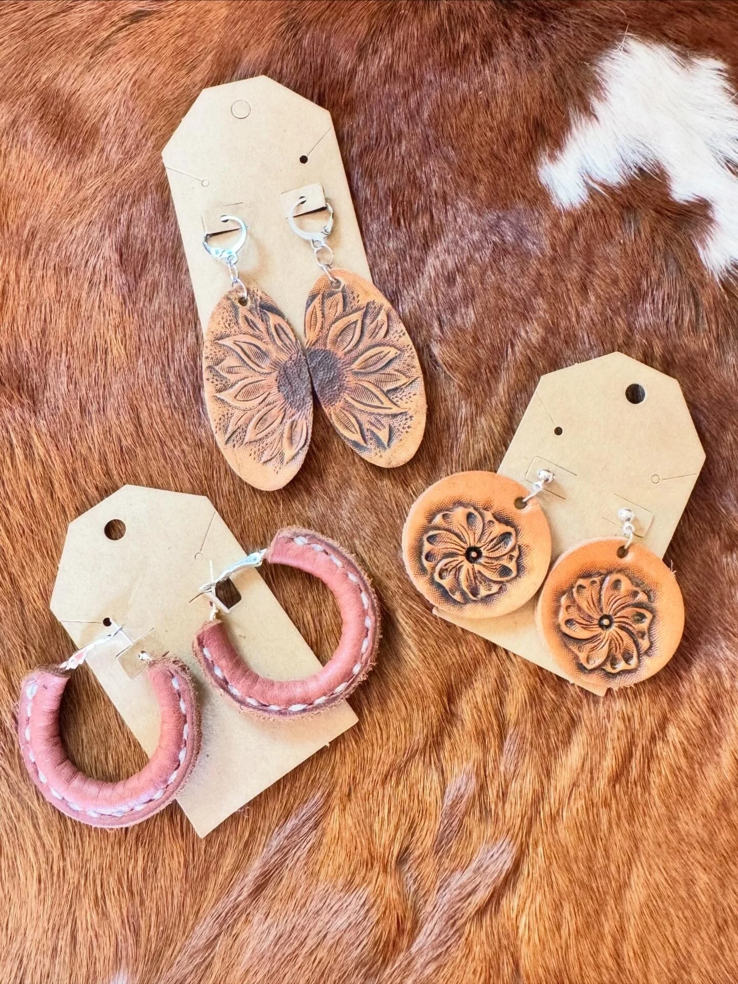 A pair of earrings featuring butterfly-shaped leather designs with intricate patterns, displayed on brown paper tags. The background is a reddish-brown fur surface.