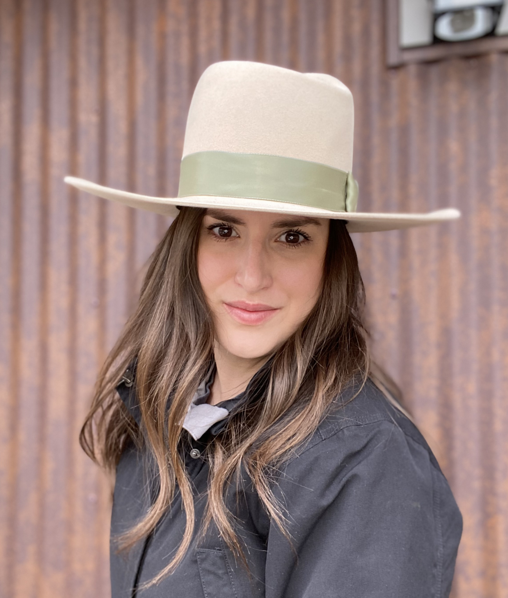 Close-up of a woman with brown hair wearing a beige hat with a green band, black jacket, standing in front of a brown curtain.