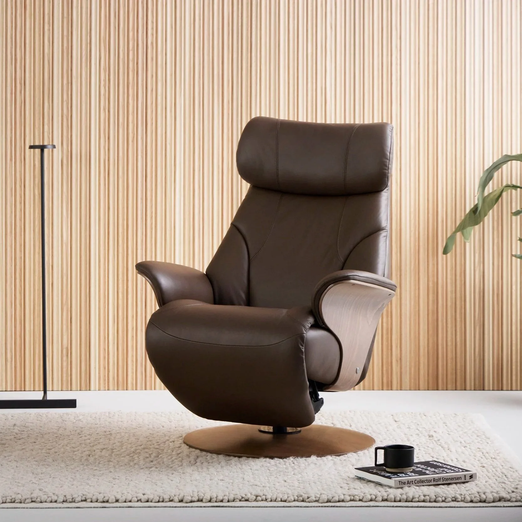 A brown leather armchair with a high back and wooden swivel base, placed on a white textured rug. To the right of the chair, a potted plant with green leaves is partially visible. To the left, a black floor lamp with a round arm. On the floor in front of the chair, there is a black mug, a closed book titled "The Art Collector Rolf Stenersen," and a small black object, all on the rug. The background features a light-colored wood-paneled wall.