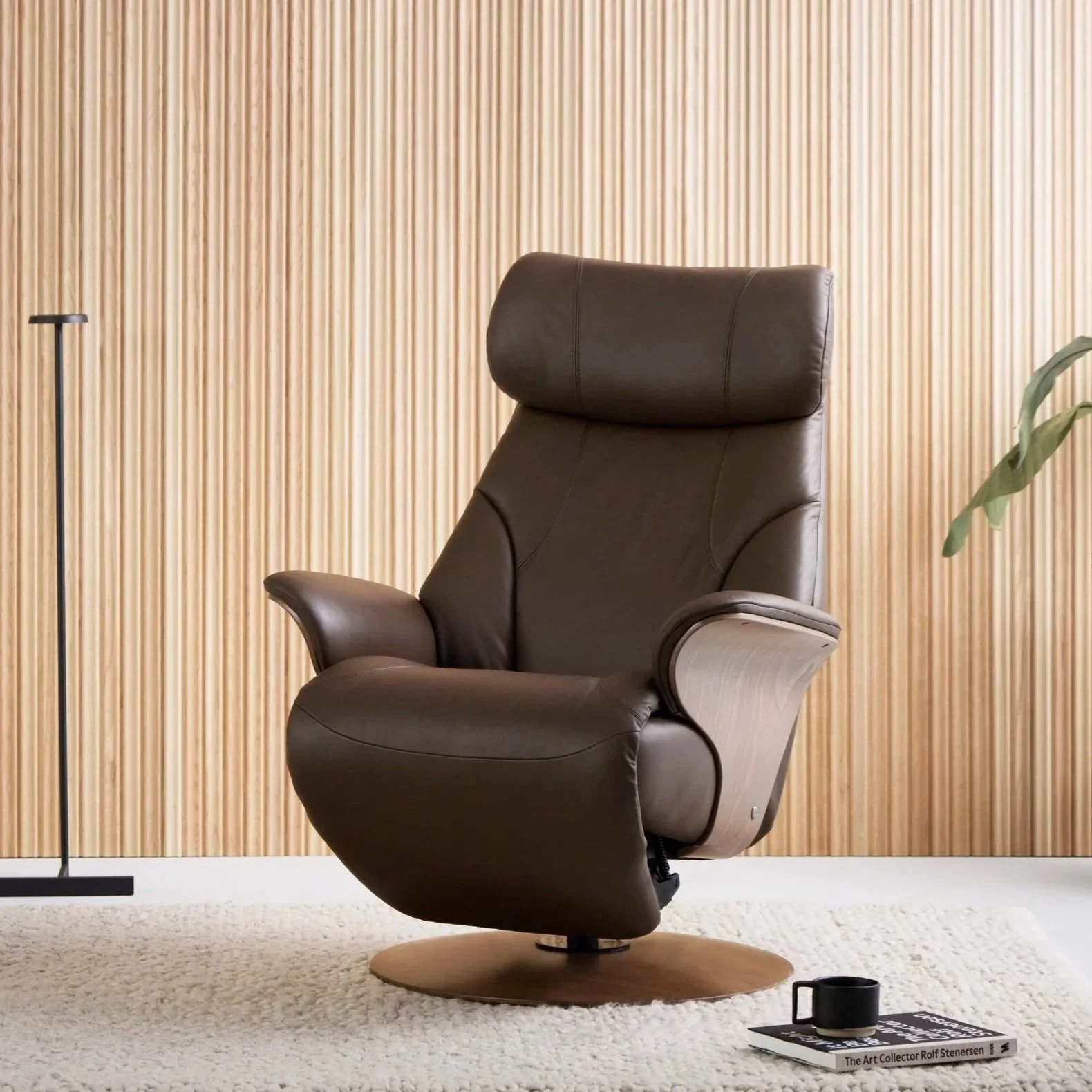 A modern, brown leather recliner chair with a headrest, set on a beige rug in front of a wooden-paneled wall. To the left, there is a black floor lamp, and on the right, a green plant. On the floor in front of the chair, there are two books, one titled "The Art Collector Rolf Stenersen," with a black coffee mug on top of them.
