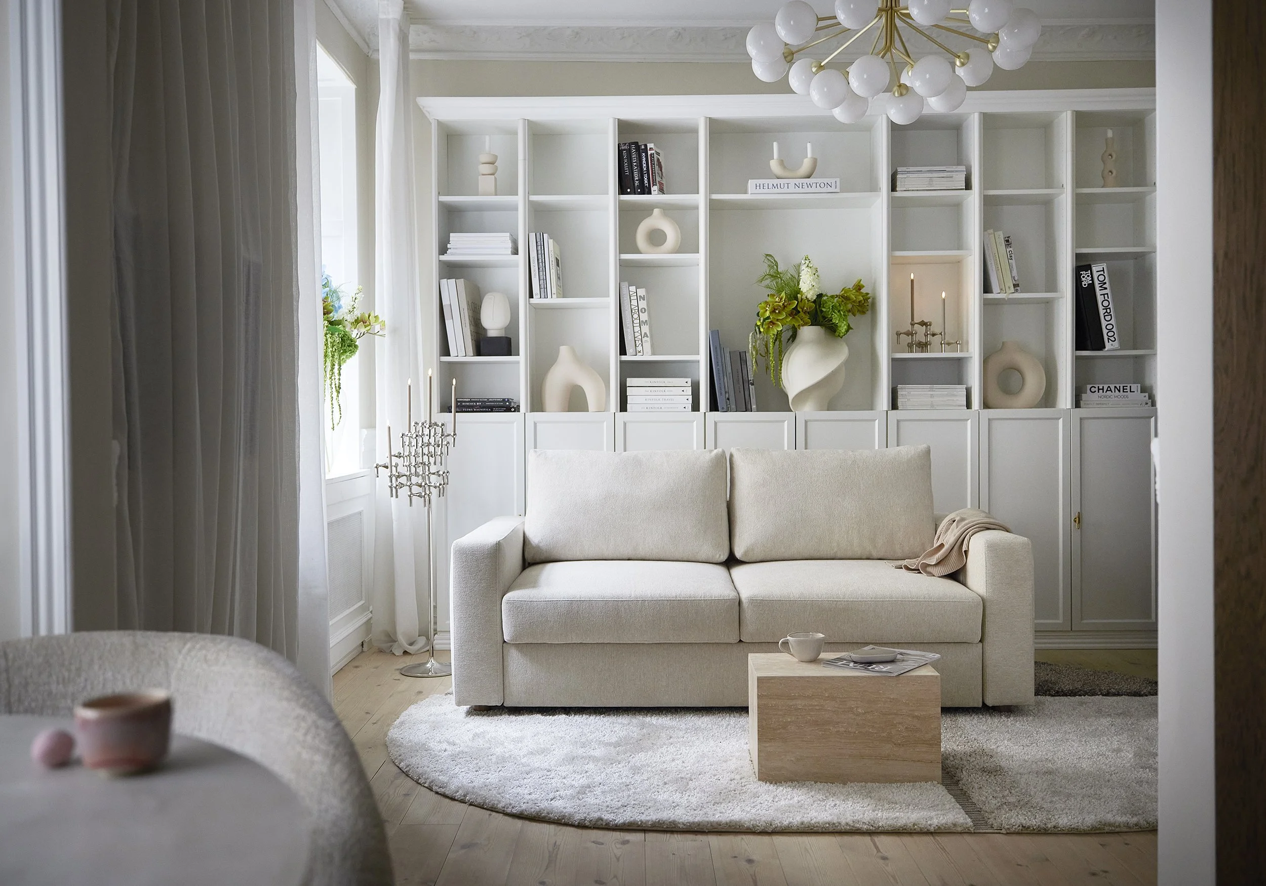 Living room with white bookshelf, beige sofa, round white rug, wooden side table, plants, modern chandelier