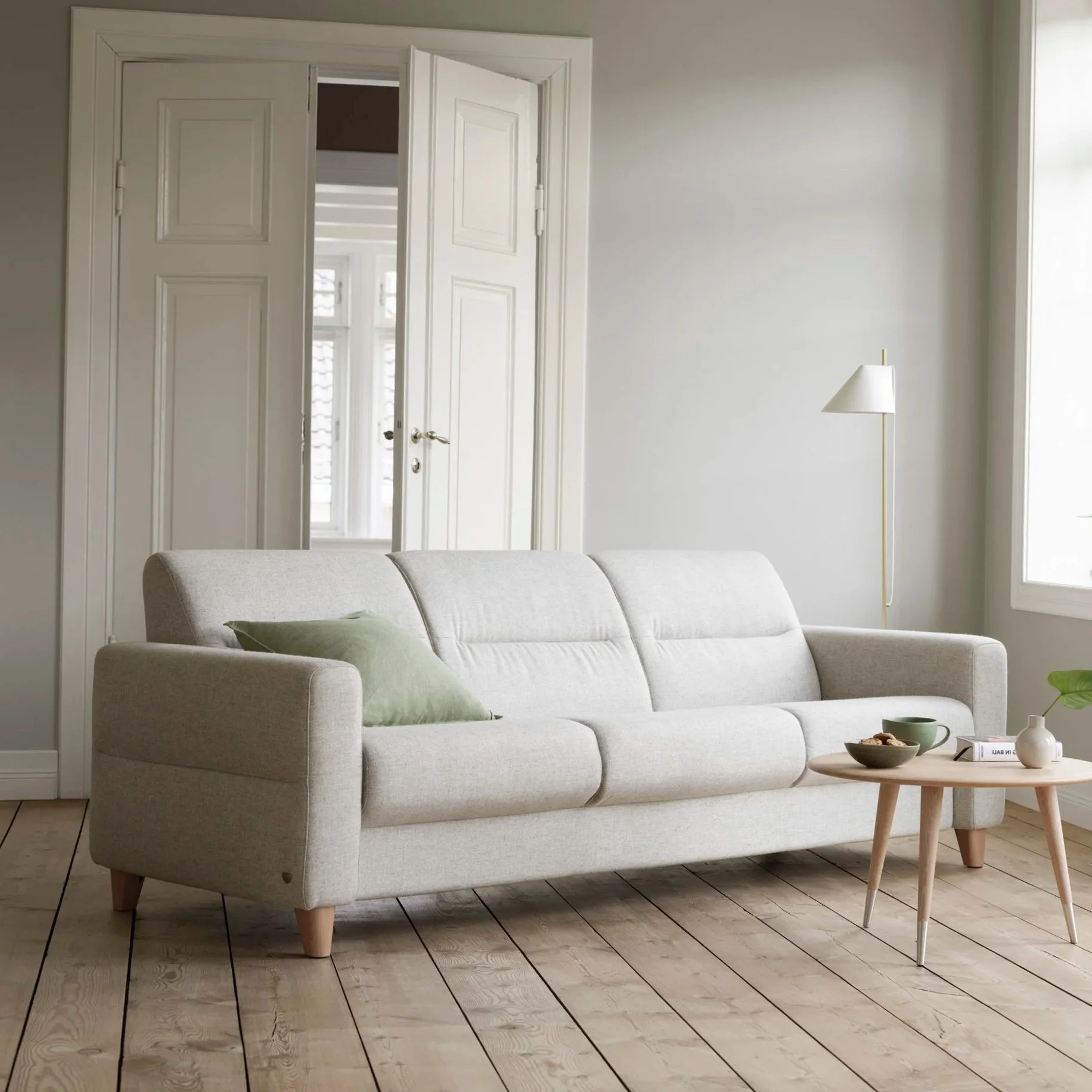 A cozy living room with a silver sofa, a green cushion, a light wood coffee table with decorative bowls and a book, a white floor lamp, and white walls with open double doors leading to a bright room with windows.